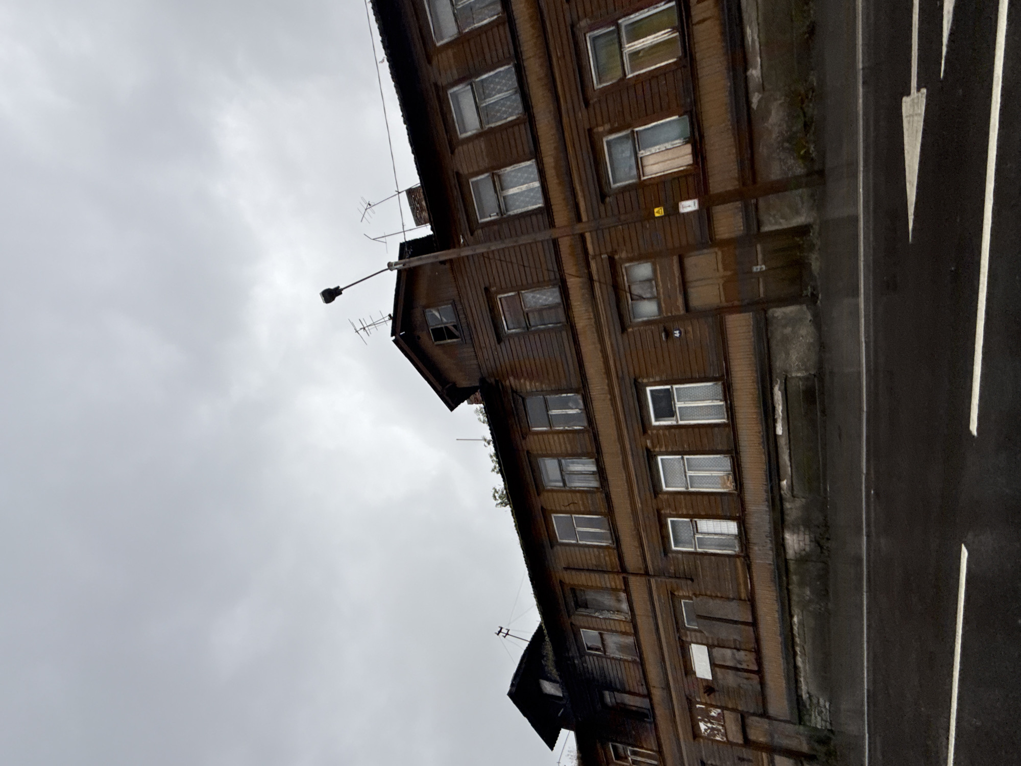 The image shows a two-story wooden apartment building with a dark brown facade and a dark, overcast sky. The building appears old and somewhat neglected, with several windows boarded up or covered. The lower level of the building has a darker, more modern-looking foundation. A street light pole stands in front of the building, and the street in front is paved and has white directional arrows painted on it. The building is located in Tallinn, Estonia. The overall impression is one of a somber, somewhat dilapidated urban scene.
