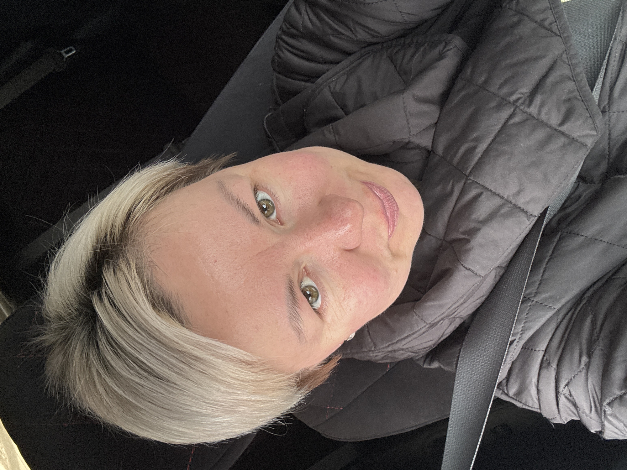 A middle-aged woman with light blonde hair is positioned in a vehicle. She's wearing a quilted jacket and a seatbelt. The interior of the vehicle is dark, with black upholstery visible. Her head is tilted, and she's looking directly at the camera with green eyes. The location of Iiul'skoe, Russia is not identifiable within the image.