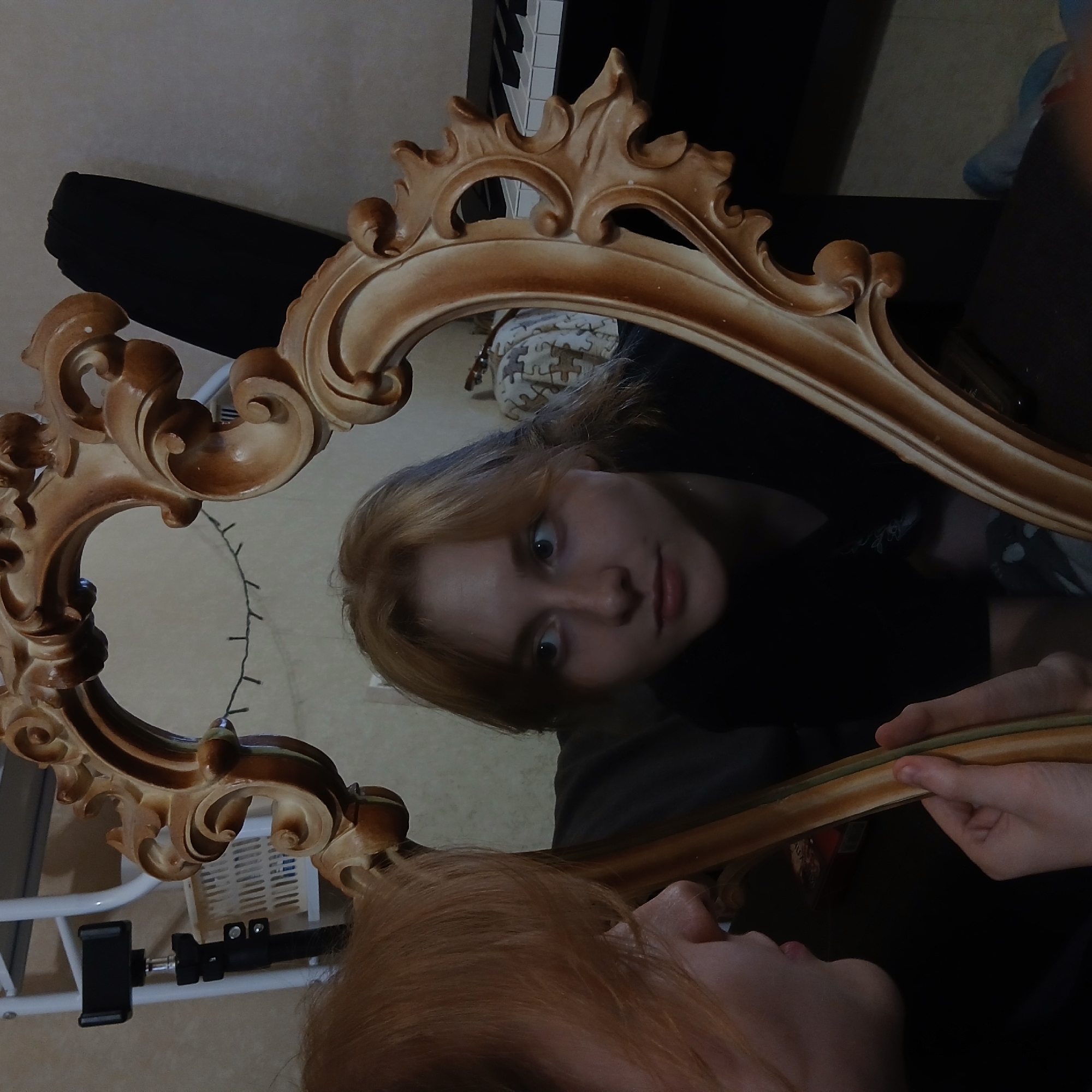 This is an indoor, self-portrait style photograph taken in Yekaterinburg, Russia, featuring a young person looking into an ornate, gilded mirror.
The primary subject is a young individual, likely female, with light reddish-blonde hair. Their face is captured in the reflection of the mirror, showing wide, light-colored eyes that convey a sense of curiosity or slight surprise, and a neutral expression. They are wearing a dark-colored top. In the foreground, outside the mirror frame, a portion of the person's chin and their hair are visible, indicating they are holding the mirror themselves. Their hands are seen at the bottom right, grasping the decorative frame.
The mirror itself is a prominent object, featuring a richly carved, scrolled frame with a warm, antique gold or bronze finish. The reflection within the mirror provides glimpses of the surrounding room: a section of a black and white piano keyboard is visible towards the top left, a patterned fabric or cushion adorned with puzzle pieces appears behind the reflected head, and a string of fairy lights hangs loosely. Further down, a white gridded object, possibly an air vent or heater, and a black phone holder attached to a stand are also reflected.
The image suggests a casual moment, possibly taken by the subject themselves, exploring their reflection or composing a photograph. There is no visible text in the image.