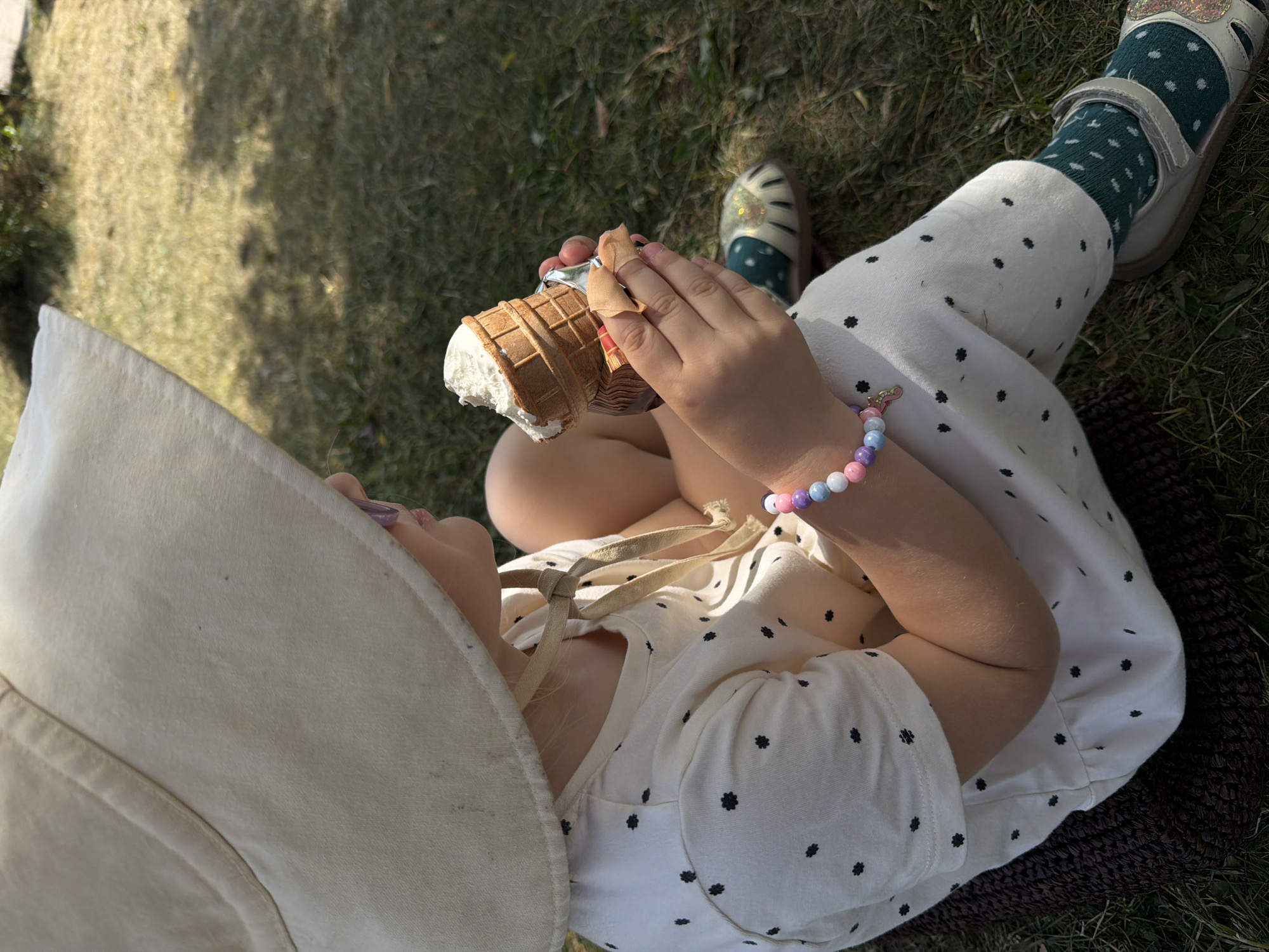 A young child is seated outdoors on a dark brown woven mat on sun-dappled grass in Khimki, Russia, viewed from a diagonal upper perspective. The child's face is largely obscured by a wide-brimmed, light beige fabric hat with ties knotted under the chin; only the right cheek, mouth, and chin are partially visible. The hat exhibits minor dark smudges on the brim. The child wears a light-colored (white or cream) outfit, possibly a jumpsuit or dress, patterned with small, dark floral or star-shaped polka dots and short sleeves. On the right wrist, a multi-colored beaded bracelet in pastel shades of pink, purple, blue, and white is visible, featuring a small, light-colored charm. Both hands hold a partially eaten waffle cone containing a scoop of light-colored ice cream, likely vanilla. The base of the cone is wrapped in a silver foil-like material, and a small, light brown fabric or paper napkin is held around it to catch drips. The visible right foot wears a light-colored sandal with a glittery overlay on the toe area and a dark green sock adorned with smaller light-colored polka dots. The surrounding environment is a grassy field, with areas of dry, yellowish-brown grass interspersed with patches of greener growth, illuminated by bright sunlight that casts distinct shadows. In the blurred background, distant green foliage is discernible.