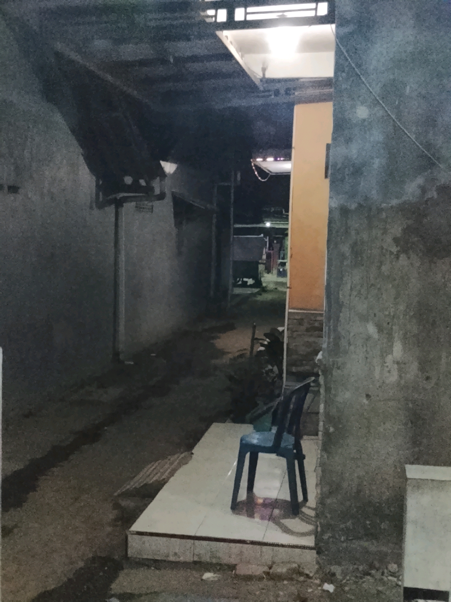 The image depicts an alleyway in Ujungrusi, Indonesia, at night. A single blue plastic chair is placed on a tiled platform in the foreground. The alley is flanked by concrete walls, with a dark tarp extending over a portion of the left side. Lights are visible in the distance, illuminating the pathway. A motorcycle is parked further down the alley. The ground is dirt and appears damp in places, with shadows cast by the overhead structures. The overall atmosphere is dim and utilitarian.
