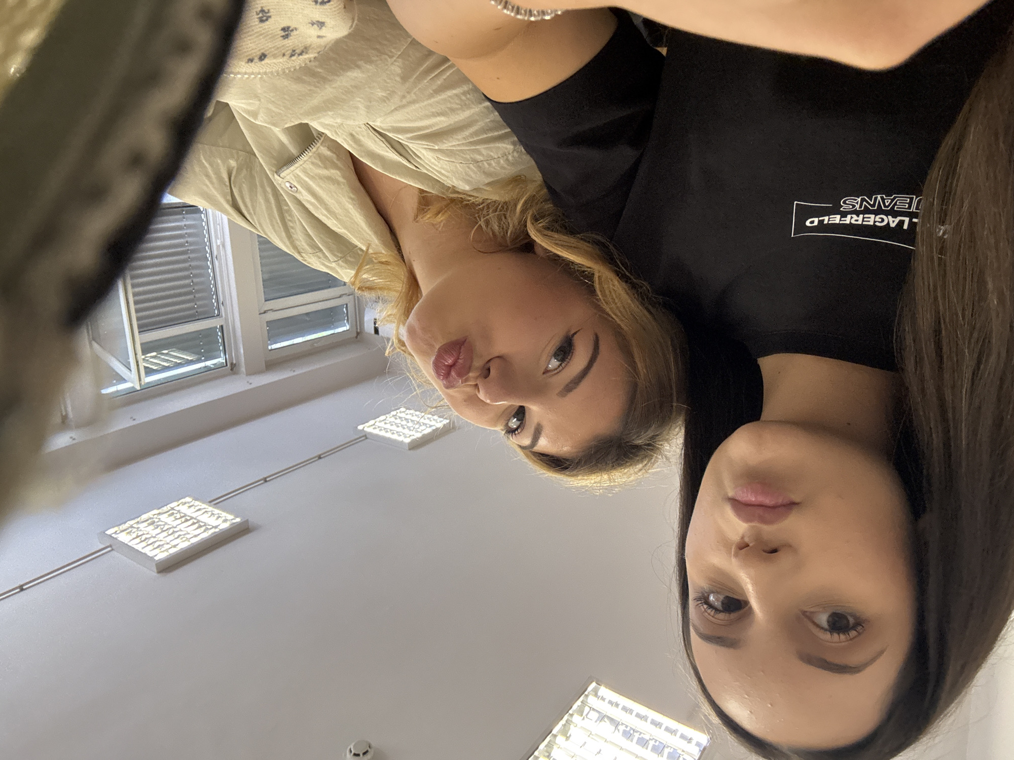 Two young women are captured in a low-angle shot, looking down towards the camera, with the image rotated approximately 90 degrees clockwise. The woman on the left has blonde, wavy hair and is wearing a light-colored, possibly beige or cream, collared top or jacket. She is making a kissy face with pursed lips and has winged eyeliner. A silver beaded bracelet is visible on her wrist.
The woman on the right has long, dark brown hair and is wearing a black t-shirt. She has a more neutral expression, looking directly at the camera. Her eyes are dark, and her eyebrows are well-defined. On her black t-shirt, the text "KARL LAGERFELD JEANS" is visible within a rectangular outline, but it appears upside down due to the image rotation.
The background shows an indoor setting with white walls and a ceiling. To the left, two windows with horizontal blinds are visible, letting in some natural light. Rectangular ceiling lights, possibly fluorescent panels, are evenly spaced across the ceiling. One light is prominently featured in the lower center of the frame, and another is visible further back. A small, round object, possibly a smoke detector or fire alarm, is also visible on the ceiling in the lower left quadrant. The overall impression is that the photo was taken in a commercial or institutional building, such as an office or a school, given the windows and lighting fixtures.