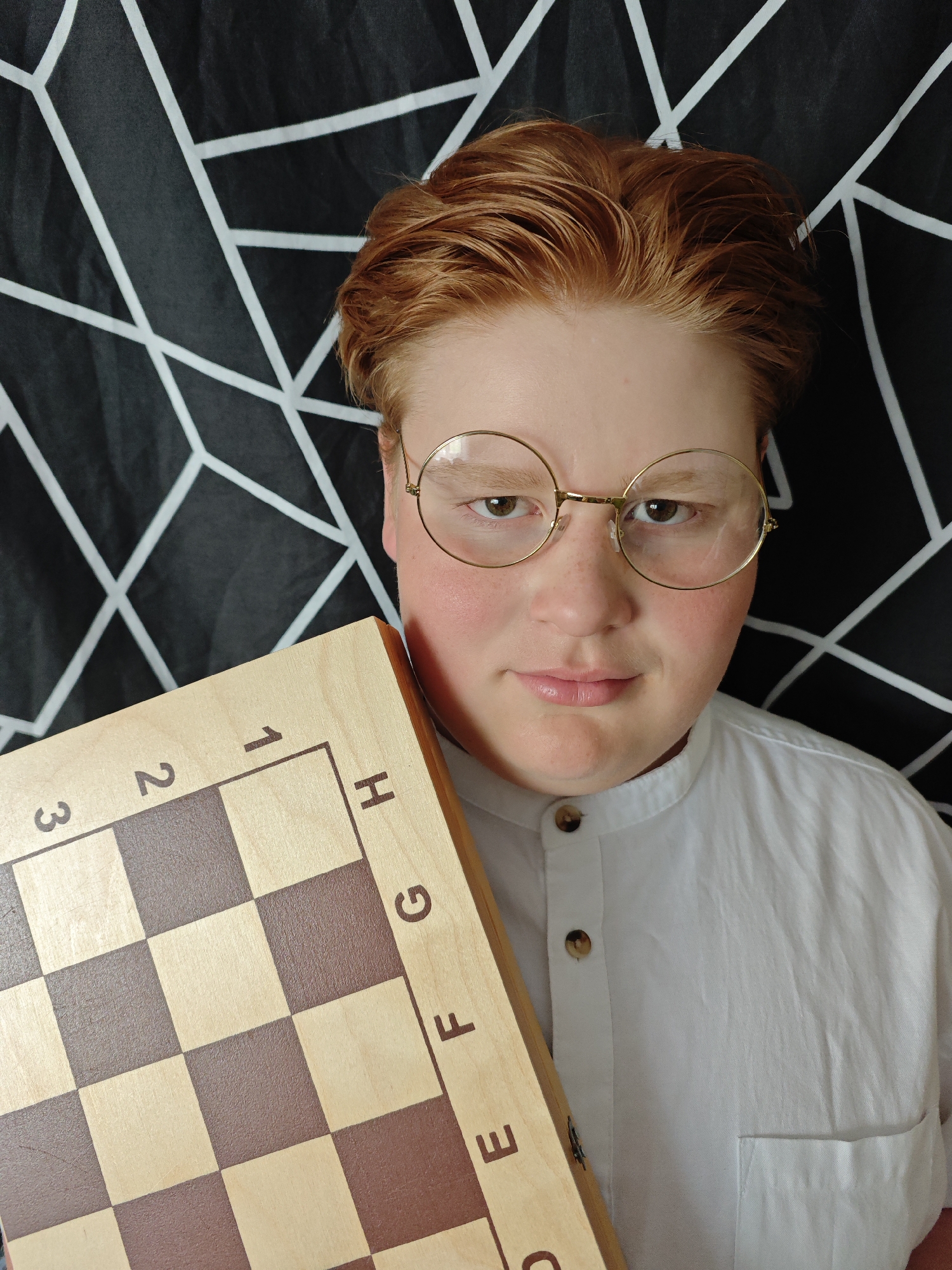 A close-up shot captures a person holding a chess board. The individual, wearing round glasses and a white button-down shirt, faces the camera directly. They have fair skin and reddish hair. The chess board, partially obscuring the left side of the frame, displays alphanumeric markings along its edge. The background consists of a black and white geometric pattern. The setting appears to be indoors, however no other environmental clues give any indication of being in Марковский, Russia.