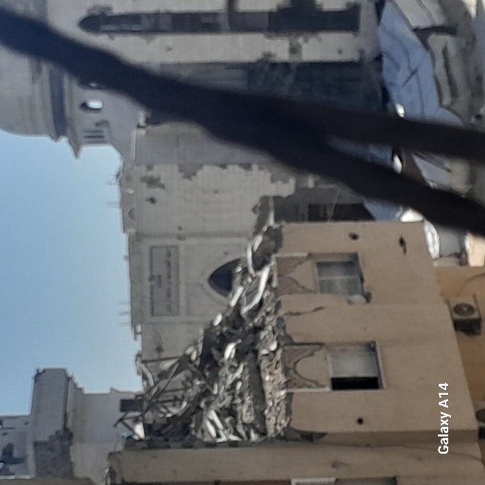 The image depicts a scene of significant structural damage in Khan Yunis, Palestinian Territory, under a clear blue sky. In the midground-left, a multi-story, light-brown building with decorative arched elements above two visible windows has sustained severe damage to its upper floor and roof, presenting a large pile of rubble, twisted metal, and shattered concrete. Two white air conditioning units are mounted on its exterior wall below the windows. In the background, a lighter-colored building with a large arched entrance or window and an illegible inscription above it exhibits extensive damage to its facade, including numerous pockmarks and holes. To its right, the upper section of a white, dome-topped structure, possibly a mosque, and an adjacent minaret-like tower are visible, also showing signs of wear or damage. Below these background structures, a light-colored tarp covers an area. The foreground on the right is partially obscured by a dark, blurred, V-shaped metallic structure. No individuals are visible. The bottom-left corner of the image contains the text "Galaxy A14."