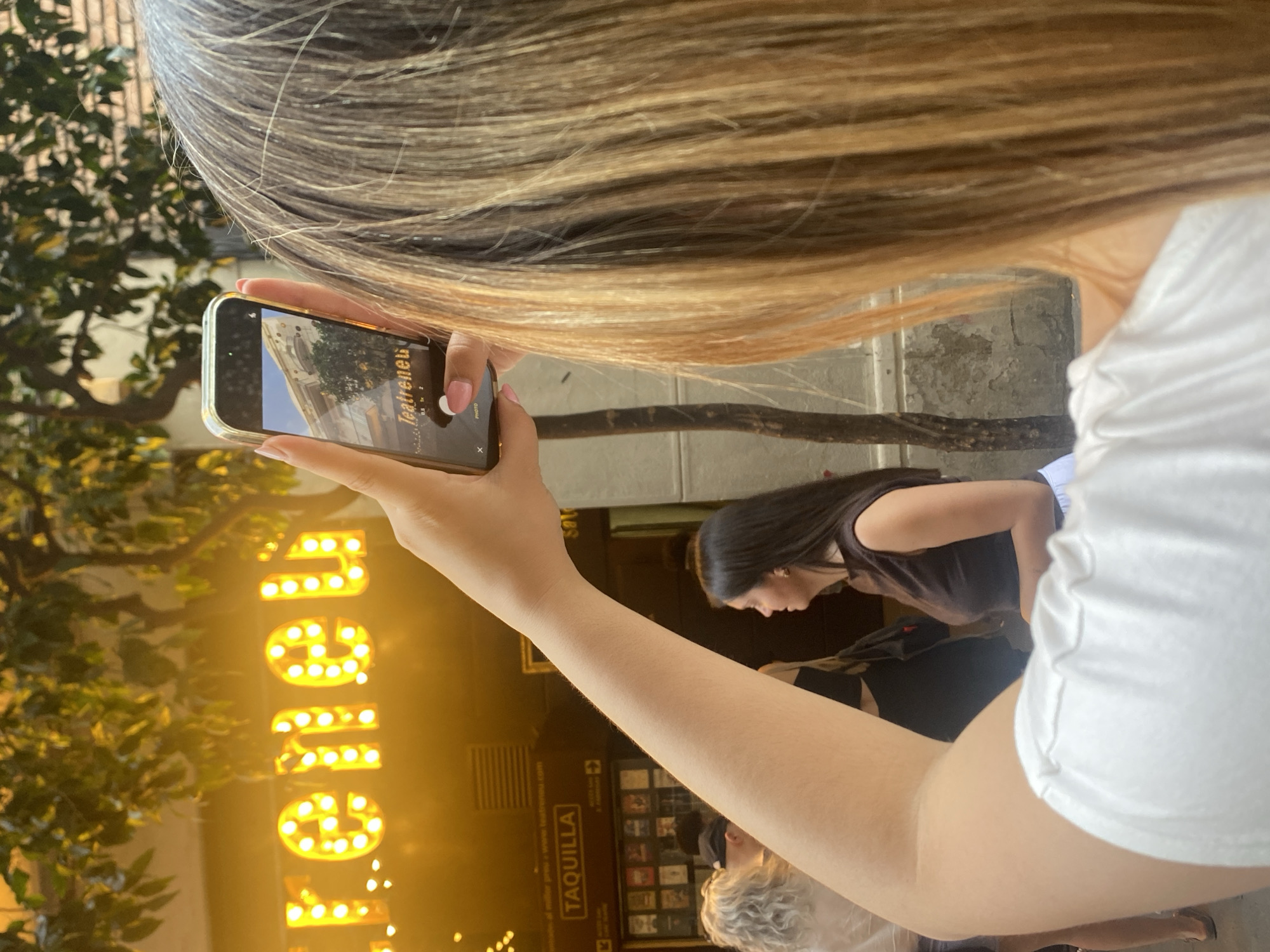 In Barcelona, Spain, a woman in the foreground, seen from the back, holds a smartphone in her right hand, angled to capture a photo of a building facade with illuminated letters. The illuminated sign reads "Teatrenen" in yellow bulbs. She wears a white shirt with ruffled sleeves. Behind her, to the left, is a ticket booth labeled "TAQUILLA" displaying various posters. Other individuals are visible in the background, including a woman with curly blonde hair and a person wearing a black cap. The scene appears to be a public area, possibly a street in front of a theater or performance venue, given the presence of the ticket booth and the marquee-like sign. A tree with green leaves extends from the upper left of the frame, casting shadows. The lighting suggests it could be late afternoon or early evening, with artificial lights from the sign contributing to the overall illumination. The architectural style of the building in the background appears to be classic, with ornate detailing.
