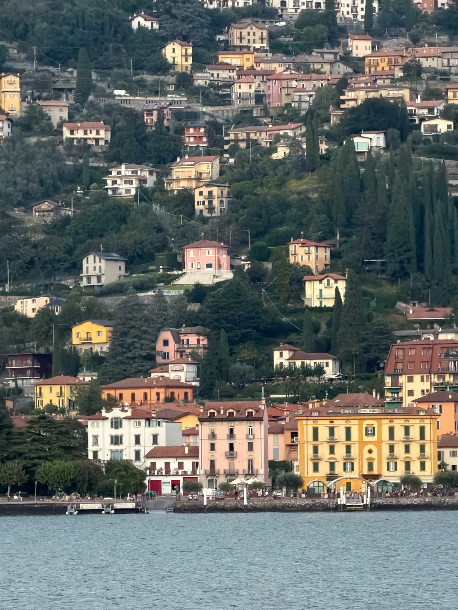 A densely populated hillside town of Varenna, Italy, is pictured from across a body of water, likely Lake Como. The town's buildings, characterized by red-tiled roofs and pastel-colored facades, are densely packed and spread across the steep incline. Many of the structures appear to be villas or apartment buildings, with a mix of architectural styles. Vegetation, primarily trees, is interspersed among the buildings, giving the impression of a town integrated with nature. In the foreground, the blue water reflects the muted colors of the sky and town. A boardwalk or promenade runs along the waterfront, with several small boats docked and a few people visible enjoying the scenery. The overall atmosphere suggests a serene, picturesque European lakeside community.