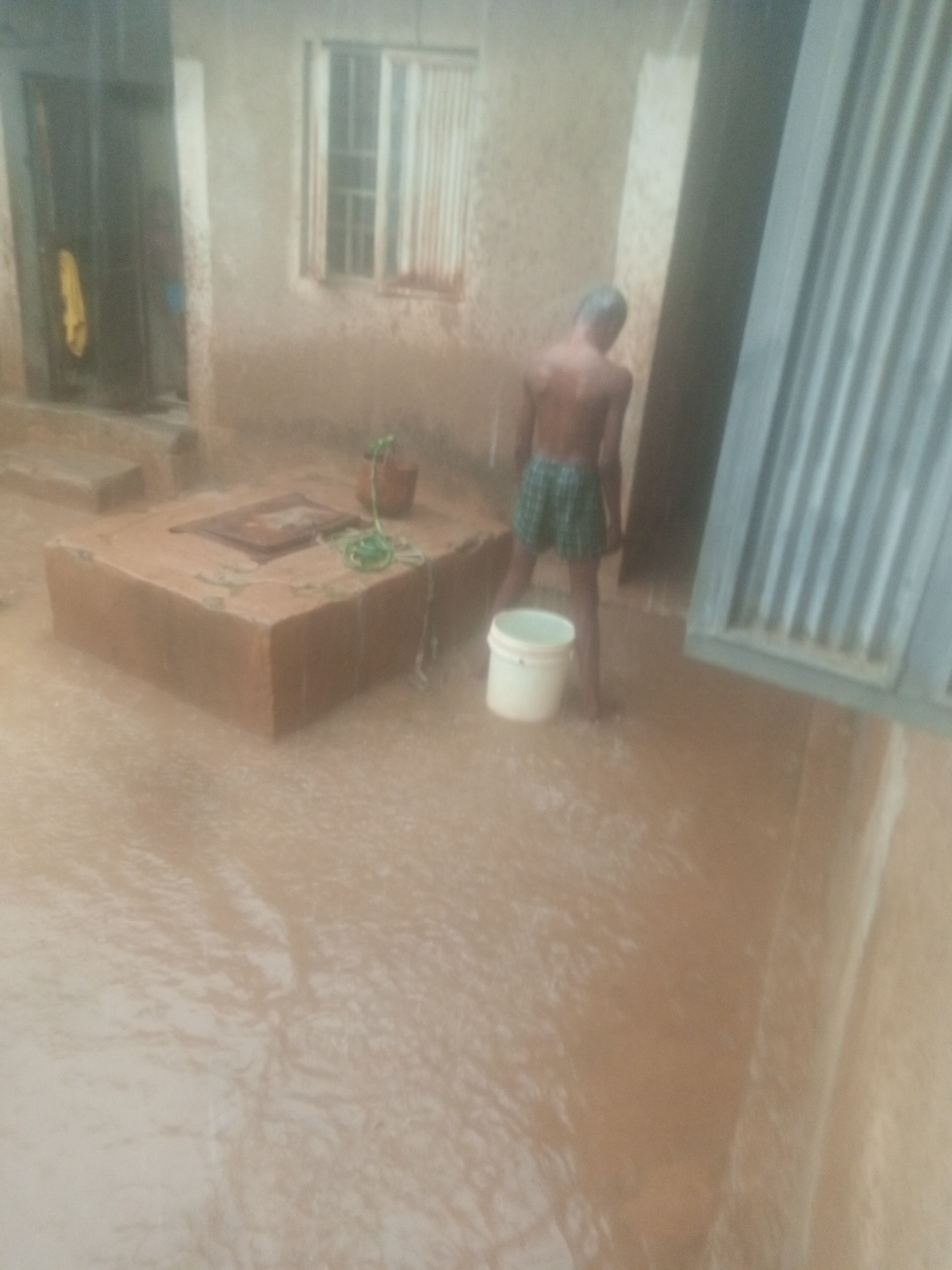 A man, shirtless and wearing green shorts, stands in floodwater with a white bucket next to him. The location appears to be the exterior of a building in Unknown, Unknown. The building is constructed of rough-textured light-colored material. There is a small window with metal bars and vertical metal siding partially visible on the right. Floodwater covers the ground, surrounding a raised, square-shaped structure made of brick or stone. A potted plant sits on this structure. There is a doorway visible on the left. It is raining.