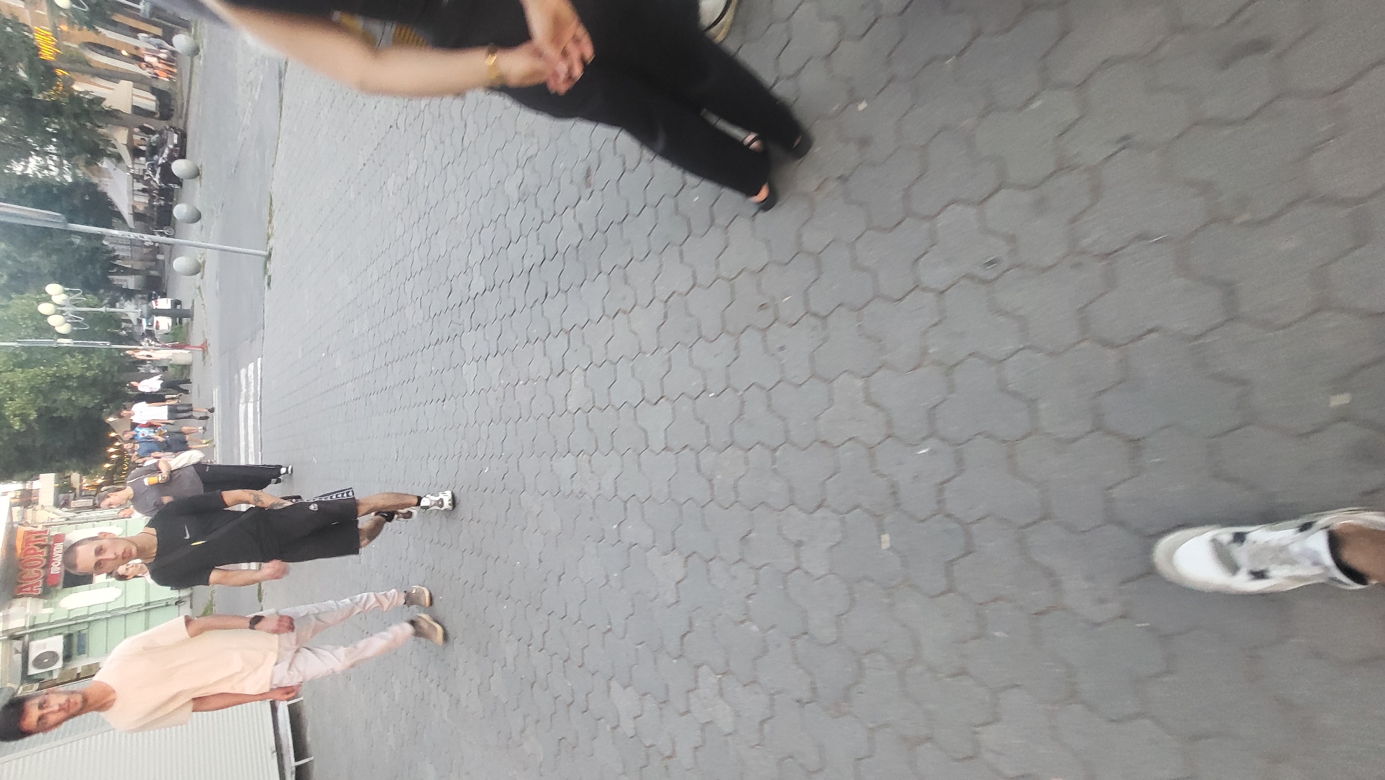 The image shows a street scene with several people. In the foreground, there are two young men. One is wearing a light-colored t-shirt and light-colored pants, and the other is wearing a black t-shirt and black shorts. They appear to be walking or standing on a paved sidewalk made of interlocking gray bricks. Further in the background, more people are visible, some walking and some standing. There are buildings and trees on the left side of the image, suggesting an urban or town setting. Streetlights with multiple bulbs are also visible. The lighting suggests it might be daytime, possibly late afternoon or early evening. There is some blurred text on a building in the background, but it is not legible. The overall scene appears to be a casual, public gathering or street activity.