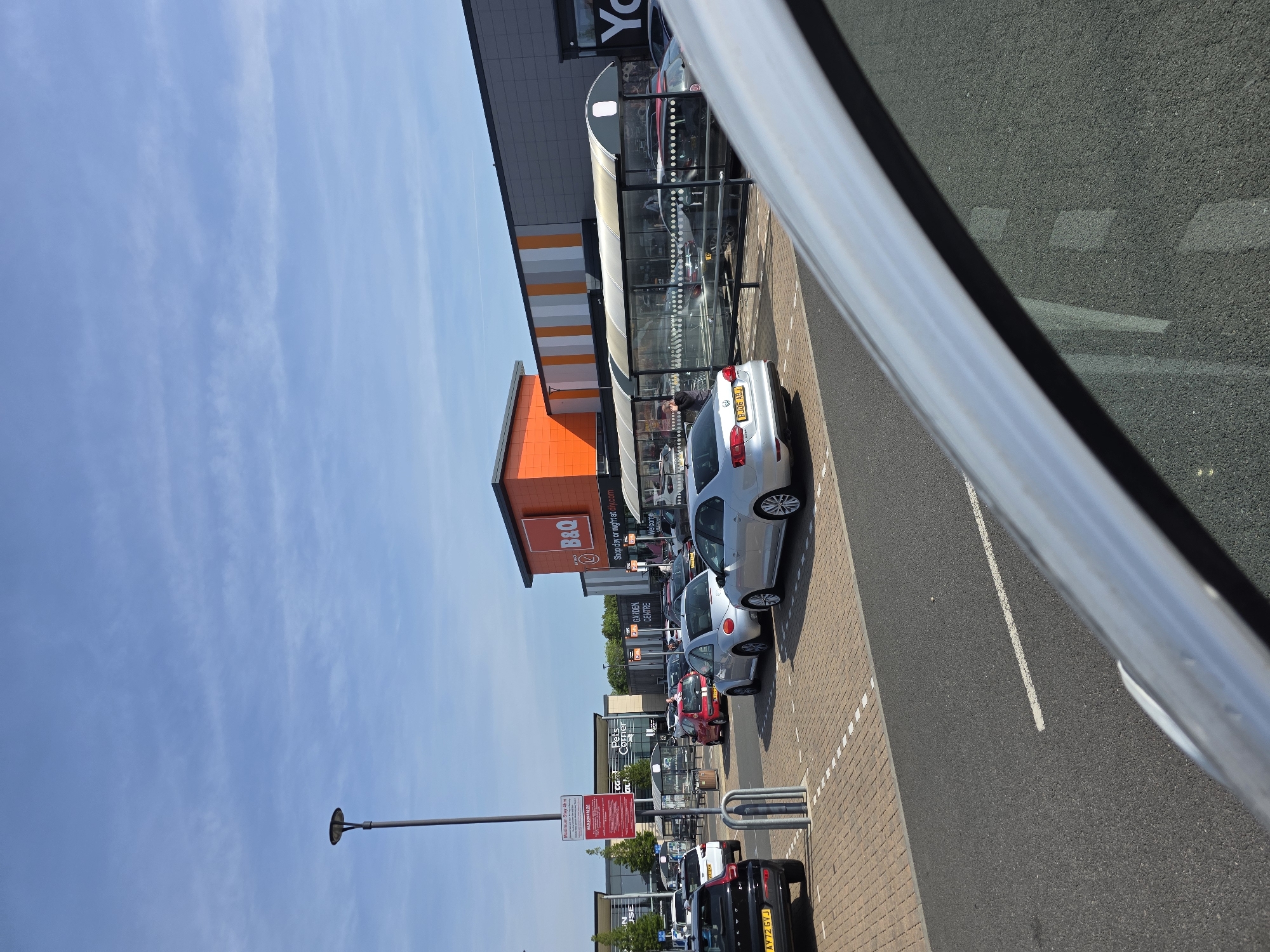 The image shows a view from inside a vehicle, looking out at a parking lot of a shopping area in Colchester, United Kingdom. Visible establishments include B&Q, Pets Corner, and an establishment labeled "Yo". Several cars are parked in designated spaces, including a silver car with license plate "BJ09 ABJ" and a black SUV with license plate "AY72 GVJ". A covered walkway runs along the front of some of the stores. There is a light pole and signs regarding parking restrictions. The sky is blue with wispy clouds.