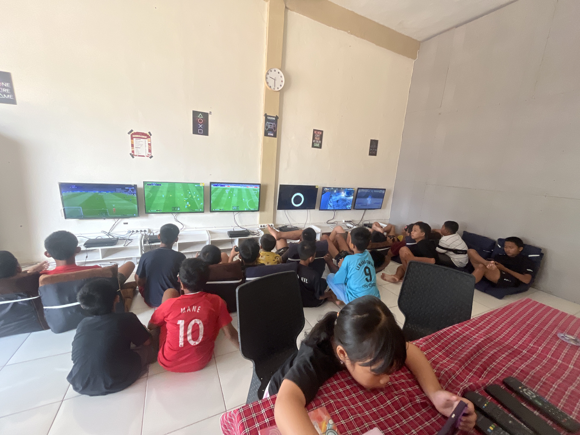 In a room in Sekayu, Indonesia, a group of children are gathered to play video games. Several televisions are mounted on the wall, displaying game content. The children are seated on the floor, facing the screens, with some using game controllers. The room appears to be a gaming center or a dedicated play area, with posters related to gaming visible on the walls, including PlayStation symbols and game-related text. The flooring is tiled. A red and white checkered blanket is spread out in the foreground, with remote controls placed on it. The overall atmosphere suggests a casual and focused gaming session.