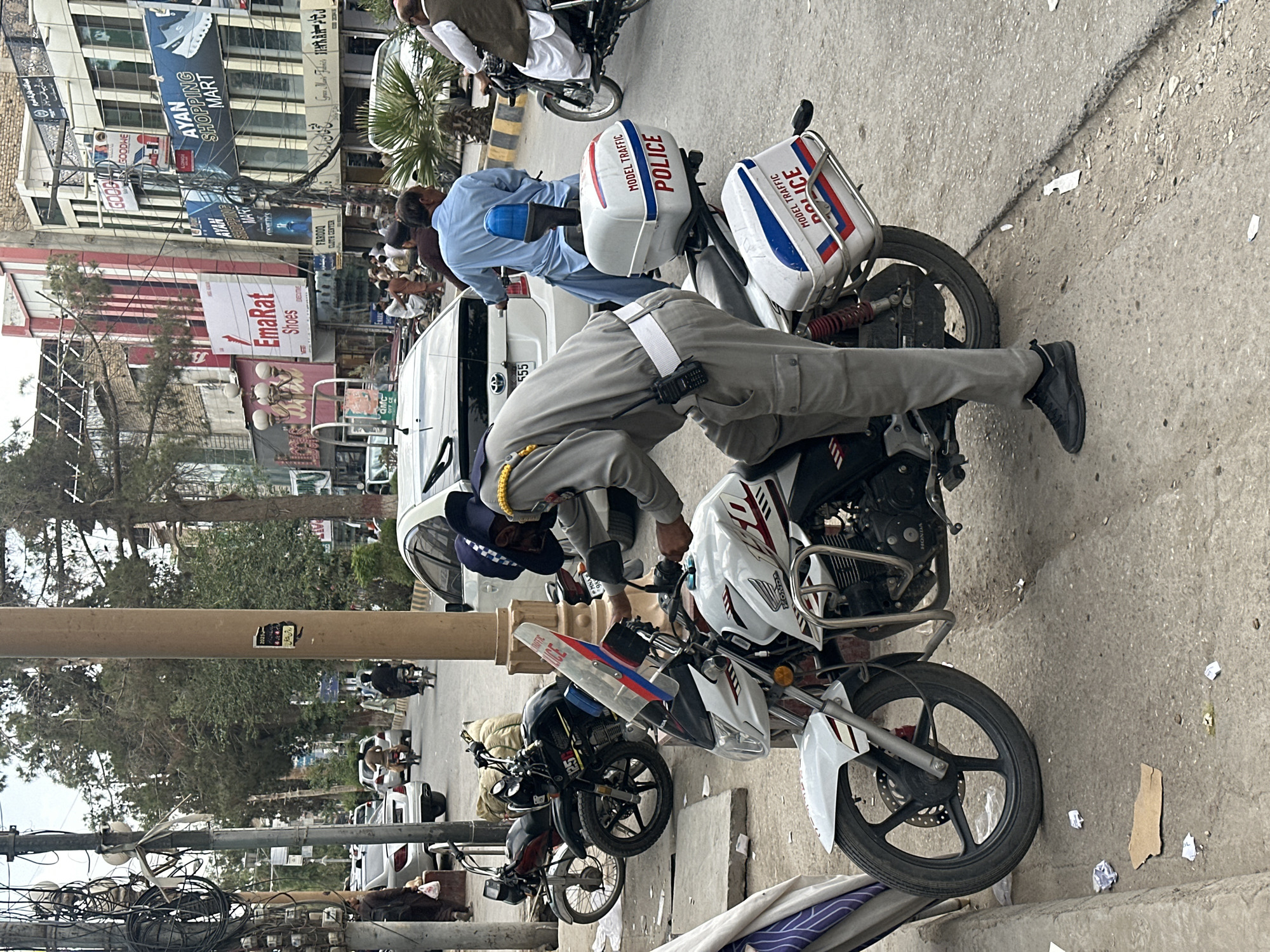 The image depicts a street scene in Quetta City Tehsil, Pakistan. A man, possibly a police officer, is inspecting a white Honda motorcycle. The street also contains several other motorcycles, some marked as "POLICE". In the background are buildings with signs such as "AYAN SHOPPING MART", "EmaRat Shoes", and "FARDOO CLOTH CENTER". There are also pedestrians visible in the background. A Toyota car with the license plate "QMC 555" can be partially seen. The electrical wiring and the utility pole with the wires are visible.
