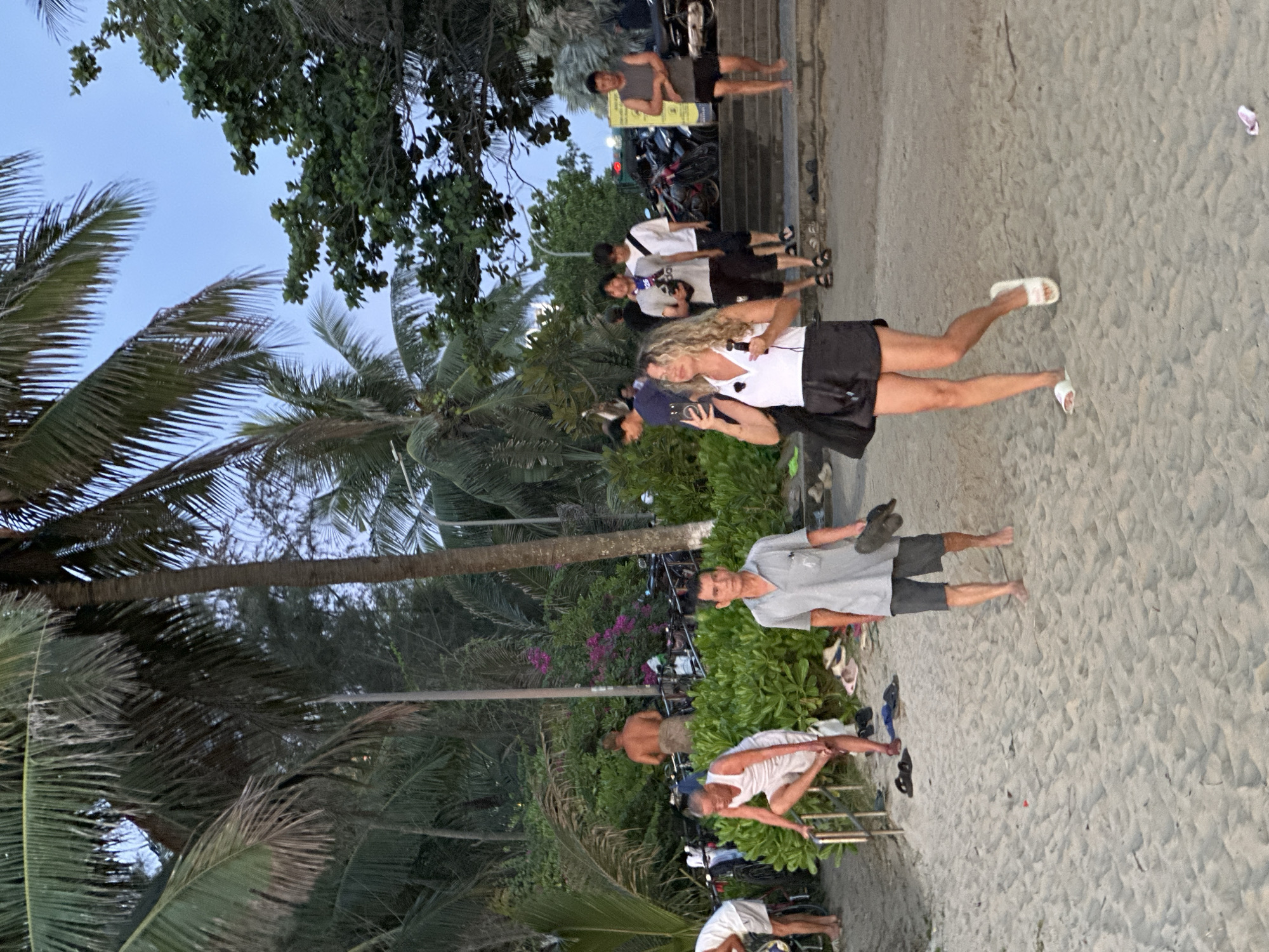 The image depicts a beach scene in Đà Nẵng, Vietnam, likely at dusk. Palm trees and lush green foliage frame the sandy beach, with a clear blue sky overhead. In the foreground, a woman with blonde curly hair, wearing a white sleeveless top and black shorts, is holding a smartphone and what appears to be a microphone, possibly recording or taking photos. She is wearing white slide sandals. To her left, a man in a grey polo shirt and dark shorts is walking, carrying dark sandals in his right hand. To the left of him, a man in a white tank top and white shorts is leaning on a metal railing, appearing to attend to his foot. Further back, several other individuals are visible, some near parked motorcycles and a stairway, suggesting proximity to a road or promenade. The overall atmosphere is casual and relaxed, consistent with a beach environment in Vietnam.