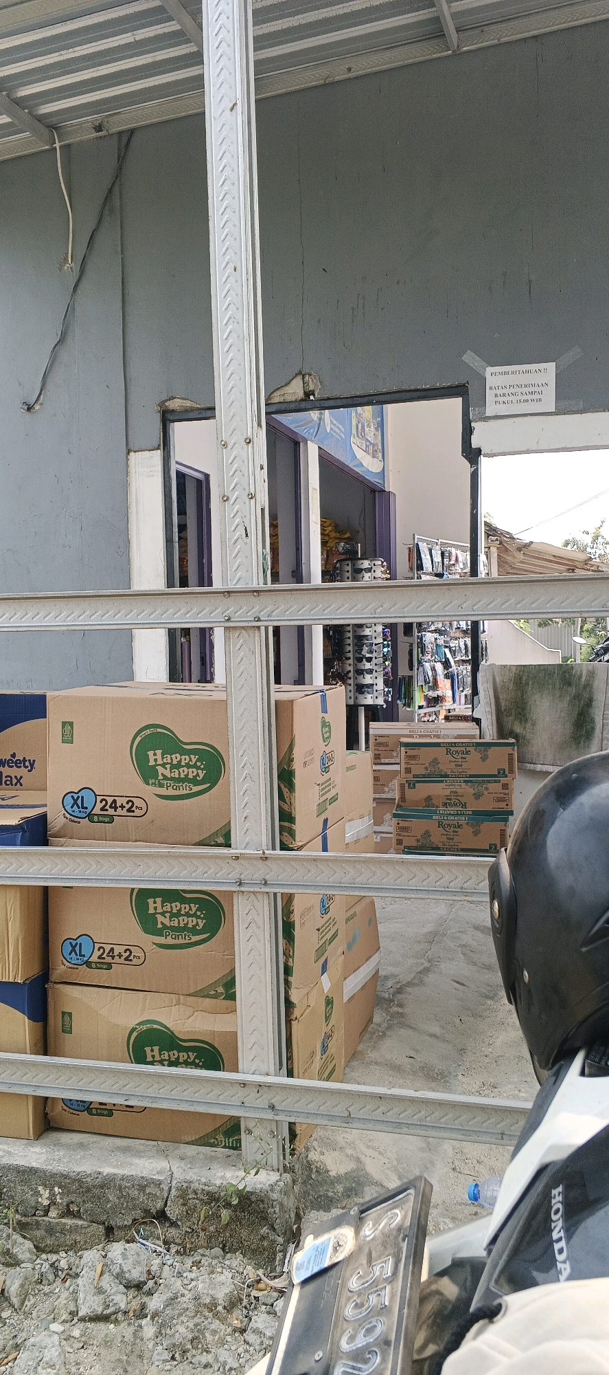 The image depicts an outdoor scene in Sonoadi, Indonesia, with a focus on stacked cardboard boxes in front of a building. The building appears to be a shop or warehouse, with a gray painted exterior and a corrugated metal roof supported by metal beams. A large, white metal pole stands vertically in the foreground, part of the structure's support system.
Several large cardboard boxes are visible, some with branding. One box prominently displays "Happy Nappy Pants" and indicates size XL with "24+2 Pcs" and "8 Bags". Another box has "sweety" and "Max" printed on it. Smaller boxes of "Royale" are also stacked.
Through an open doorway in the building, more merchandise is visible, including clothing racks with various items, sunglasses displayed on a stand, and other products on shelves. A sign is taped to the building's wall above the doorway, with text that appears to be in Indonesian, mentioning "PEMBERITAHUAN" (Notification) and "BATAS PENERIMAAN BARANG SAMPAI PUKUL 15.00 WIB" (Goods receiving limit until 15:00 WIB).
In the bottom right corner, a portion of a motorcycle is visible, including a black helmet and a license plate that reads "S 5592". A blue plastic water bottle is on the ground near the motorcycle. The ground in front of the building is concrete with some areas of debris and cracks, with a small plant growing through the concrete. The overall impression is that of a commercial establishment, likely a retail store or distribution point, with goods being stored or prepared for sale.