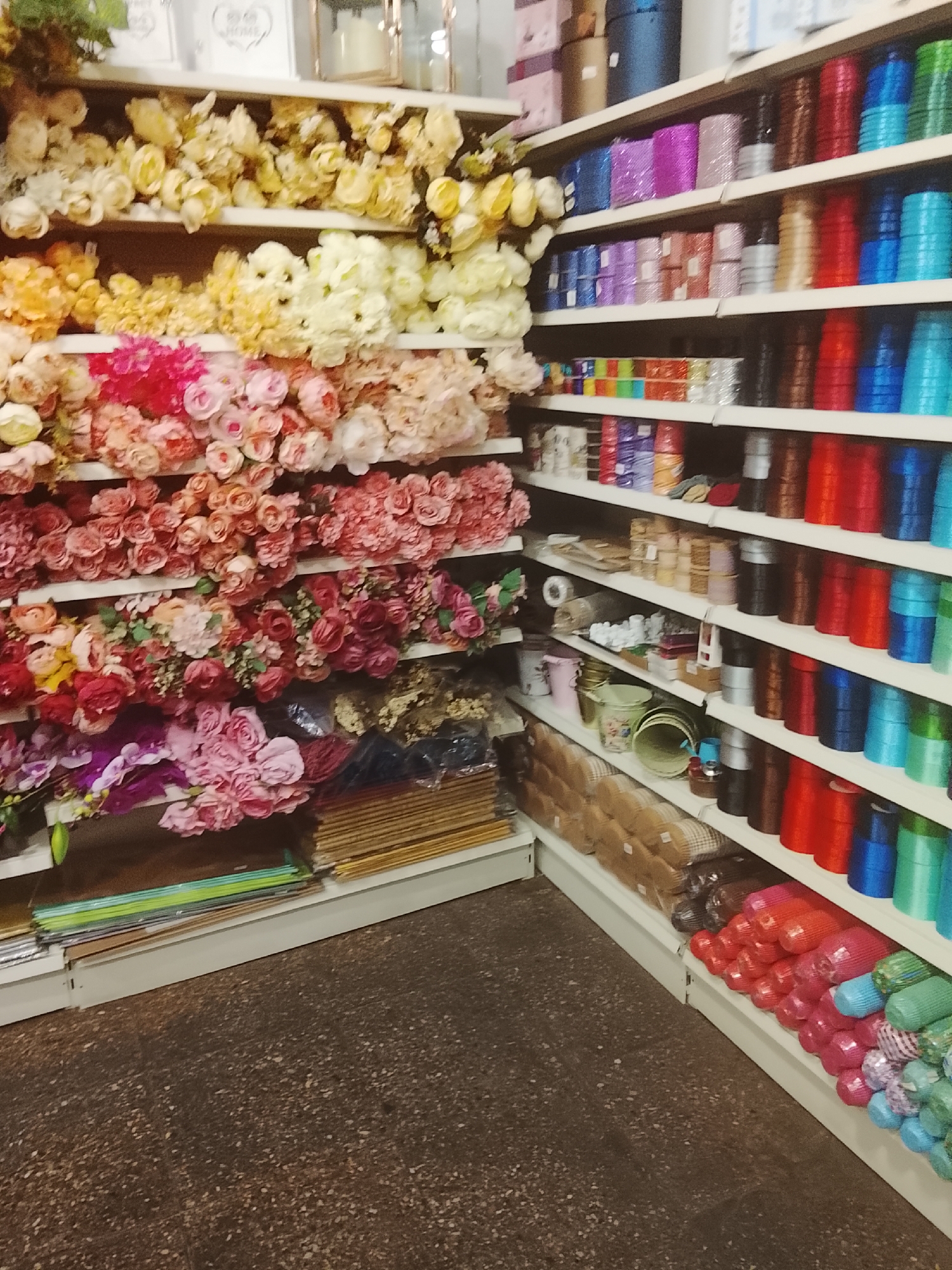 The image depicts an interior view of a retail establishment, likely a craft or hobby store, located in Unknown, Unknown. The scene is dominated by two sets of white shelving units laden with merchandise, extending from the foreground into the background on both the left and right sides of the frame.
The shelving unit on the left is primarily dedicated to artificial flowers, organized by color on multiple tiers. The upper shelves display clusters of cream, yellow, and light orange-hued flowers. Subsequent lower shelves feature dense arrangements of artificial flowers in various shades of pink and red, including numerous rose and peony-like blooms. The lowest visible shelf contains bouquets in pinks, purples, and darker reds, alongside several plastic-wrapped bundles of materials. Above the top shelf on the left, several decorative items are visible, including white framed objects, one with the word "HOME," and a metallic-toned lantern.
The shelving unit on the right displays a wide array of craft supplies, predominantly spools of ribbon and rolls of decorative mesh. The upper shelves showcase glittery ribbons and mesh in blue, magenta, pink, and silver, interspersed with solid-colored spools in brown, red, blue, and green. The middle and lower shelves are densely packed with numerous spools of solid-colored ribbon in a spectrum of hues, including red, blue, green, black, brown, and metallic tones. The bottom two visible shelves contain a large volume of individually plastic-wrapped rolls, likely ribbons or decorative wraps, organized by color, featuring items in red, pink, blue, green, and silver. A large, dark blue cylindrical box is positioned on the top edge of this shelving unit.
The floor is a dark, speckled surface, consistent with commercial or industrial flooring. No individuals are present within the captured frame.