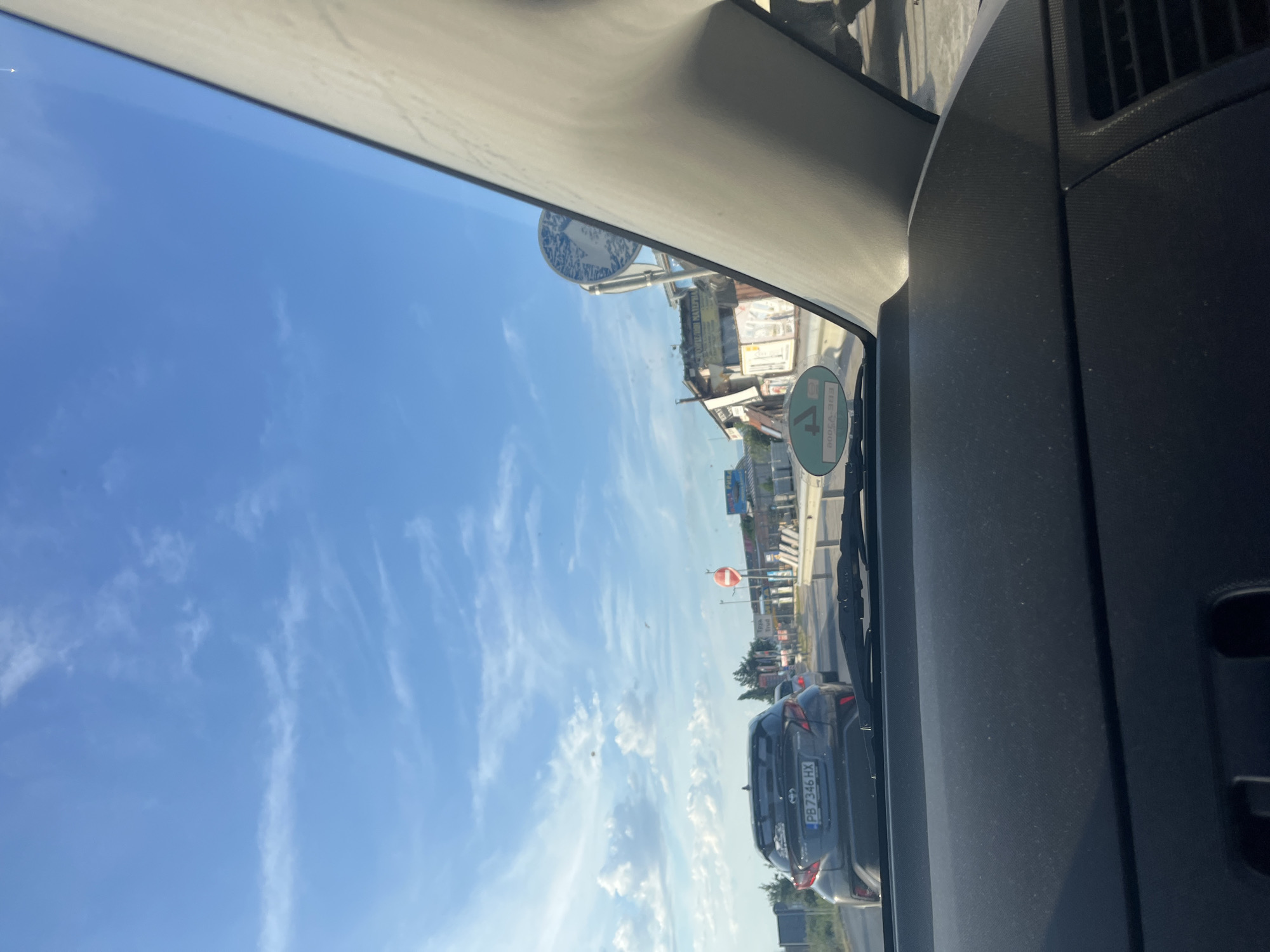 The image shows a view from inside a car, looking forward on a road. The car is in traffic, with other vehicles visible ahead. The scene is set in the city of Trud, Bulgaria. The sky is blue with scattered white clouds. On the right side of the road, there are buildings that appear to be businesses, possibly related to construction materials, with signs visible. A red and white circular traffic sign is also present. The license plate of the car in front of the viewer is visible and reads "PB 7346 HX".