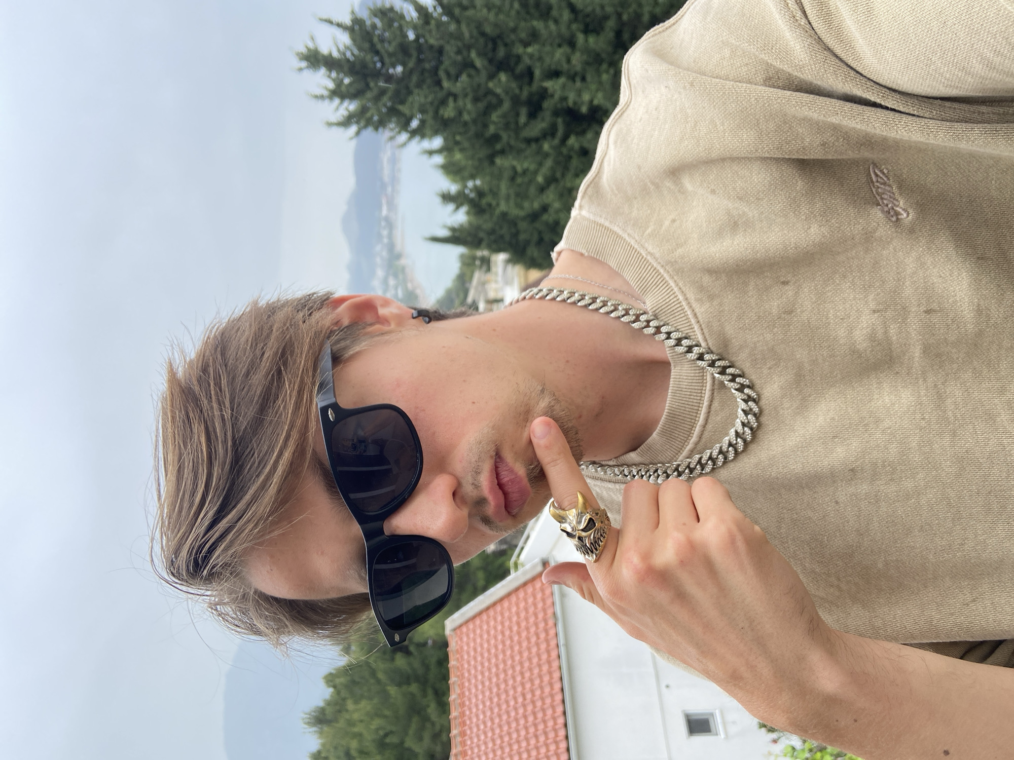 A person is posing for a photo in Šušanj, Montenegro. The person is wearing a light brown sweatshirt, a silver chain necklace, and black sunglasses. They are also wearing a large, gold-colored skull ring on their right hand, which is raised to their face. The background shows a tiled roof, a white building, green trees, the sea, and mountains in the distance. The sky is overcast.