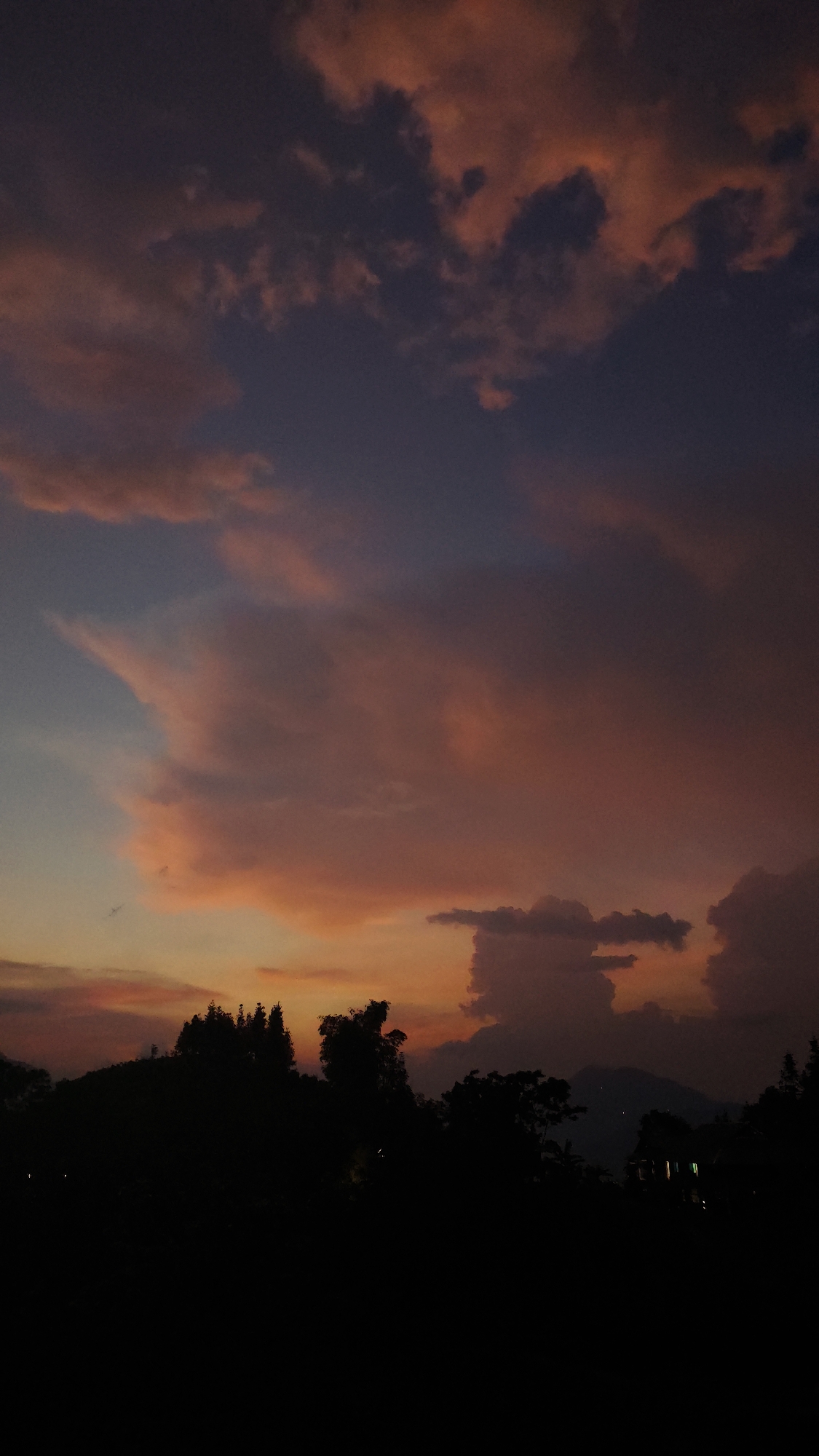 A wide shot depicts a twilight scene in Hoàng Su Phì Commune, Vietnam. The sky is filled with clouds in various shades of orange, pink, and dark blue, indicating a setting sun. Silhouetted trees and possibly mountains form the horizon line, appearing as dark masses against the colorful sky. Light sources suggest minimal habitation.