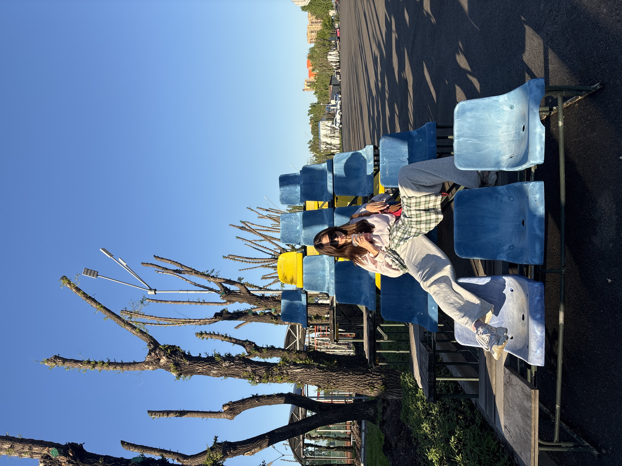 A woman is sitting on a row of blue and yellow stadium-style seats outdoors. She is wearing sunglasses, a light pink shirt, a green and white plaid shirt, and gray sweatpants, with white sneakers on her feet. The seating is located next to a paved area, with trees bordering the foreground and buildings in the background. Tall, bare trees with newly emerging green leaves are in the foreground, some equipped with street lamps. The image appears to be taken during the day under a clear blue sky. The city is Krasnoyarsk, Russia.
