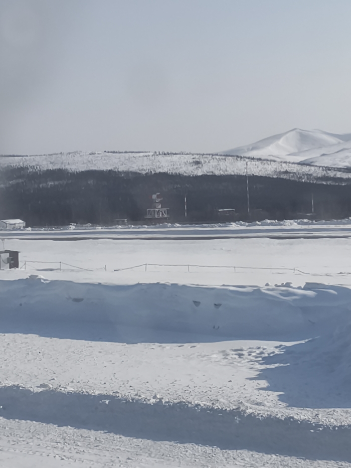 The scene depicts a snow-covered airfield environment in Sokol, Russia, under a bright, potentially hazy sky. The foreground is comprised of extensive snowdrifts and a textured snow-covered ground. Centrally, a dark, cleared strip, consistent with an airport runway or taxiway, extends horizontally across a broad, snow-laden expanse. To the left of this cleared strip, a small, dark, rectangular structure stands on the snow. In the midground, a dense line of dark evergreen trees forms a forest boundary behind the airfield. Within this tree line, a prominent lattice tower topped with a white spherical radome and multiple slender communication towers are visible. A white building, possibly an airport facility, is partially discernible on the far left within the tree line. The distant background features snow-capped mountains beneath a pale sky. No human figures or active vehicles are present in the observable area.