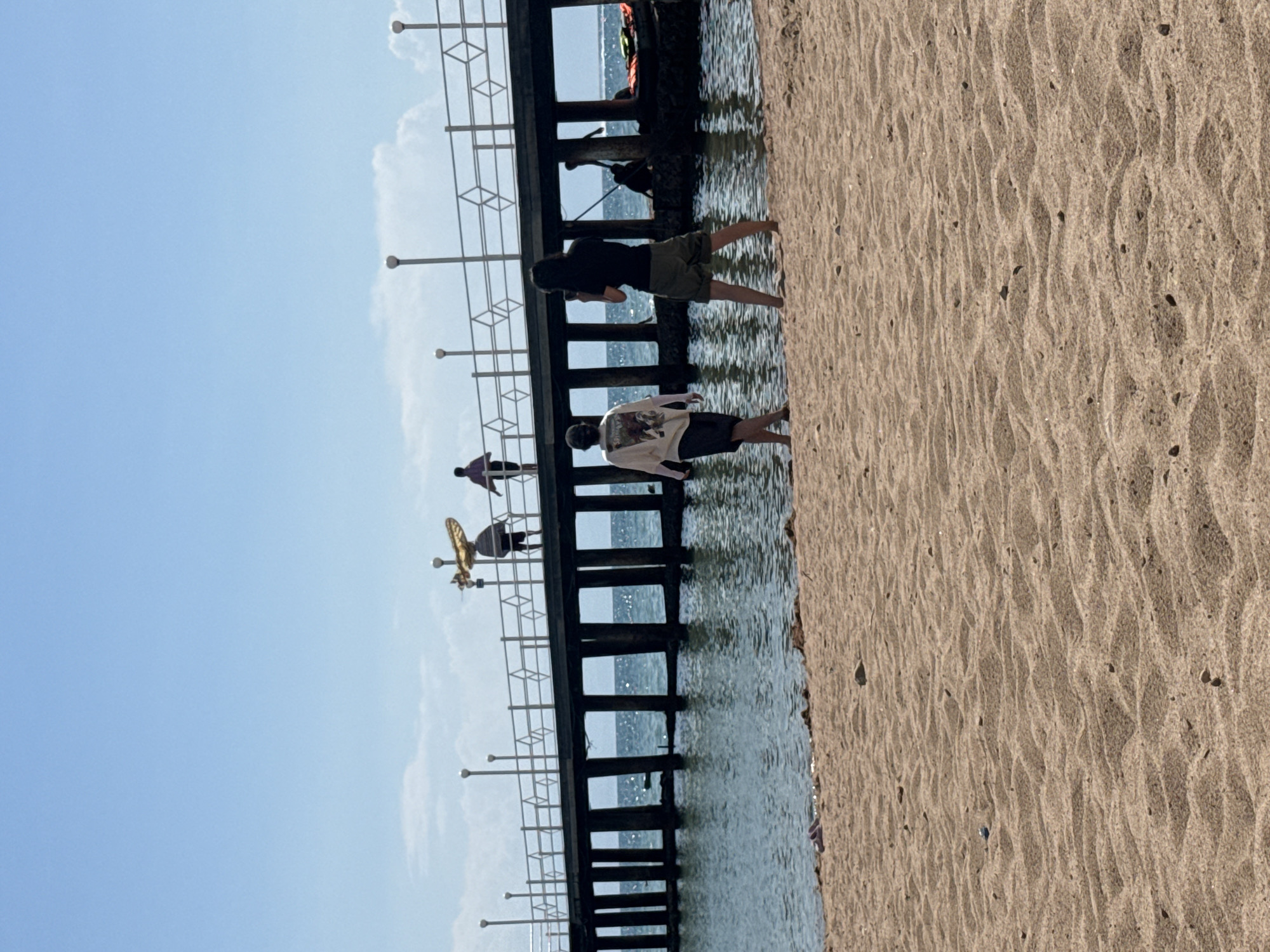The image depicts a sunny day at a beach, likely in Sary-Oy, Kyrgyzstan, with a wooden pier extending into the water. On the pier, several individuals are visible, some walking and one carrying a yellow surfboard. In the foreground, on the sandy beach, two people are standing near the water's edge. One is a person wearing a light-colored t-shirt with a graphic print and dark shorts, walking away from the camera. The other is a person wearing a black t-shirt and olive green shorts, facing towards the water and appearing to hold a phone or camera. The water is calm with ripples, and the sky is clear and blue with a few scattered clouds. The sandy beach shows patterns from the receding tide.