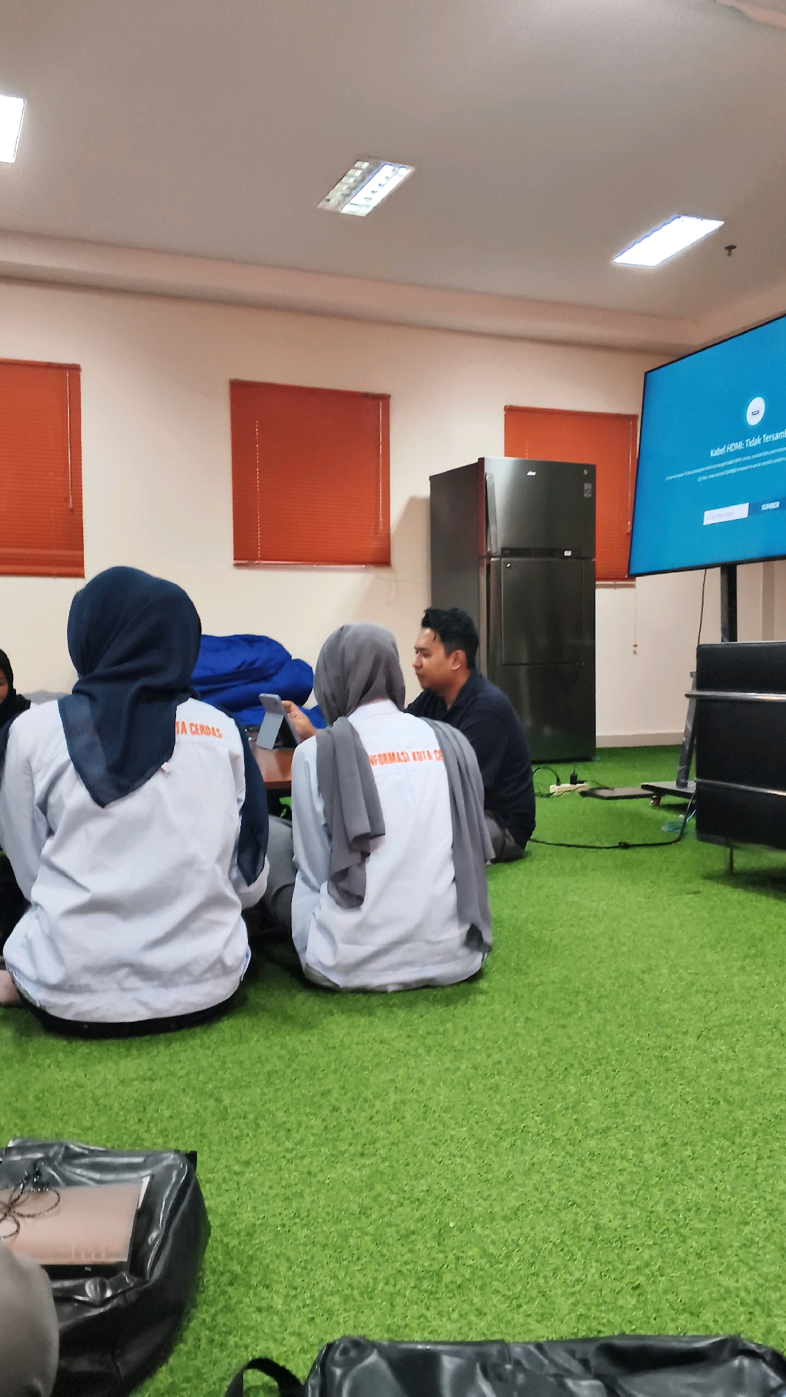 The image depicts an indoor scene with a group of people gathered on bright green artificial turf flooring, suggesting a modern, possibly informal meeting space or lounge area. The room appears to be a classroom or presentation room with a large screen mounted on the wall displaying text in Indonesian that reads "Kabel HDMI Tidak Tersambung" (HDMI Cable Not Connected). Two individuals, likely students, are seated facing away from the camera, both wearing light-colored long-sleeved shirts and headscarves. The person on the left wears a dark blue headscarf, and the person on the right wears a light grey headscarf. They are interacting with a tablet device that one of them is holding. Another person, a man wearing a dark shirt, is seated facing them, engaged in conversation. In the background, there are large orange-paned windows with blinds, a tall black refrigerator, and dark furniture. The lighting is provided by fluorescent ceiling fixtures. There are also black bags on the floor in the foreground. The overall ambiance is one of a collaborative study or work session. The location is identified as Lohbener, Indonesia, suggesting this is likely a meeting within an educational institution or business in that area.