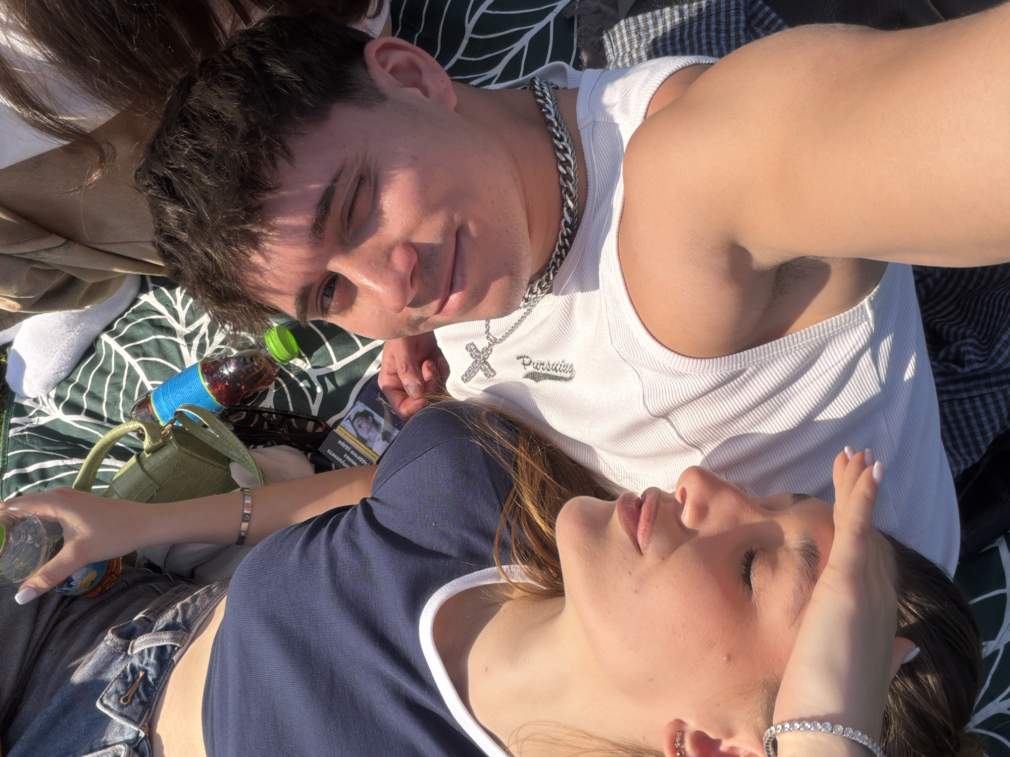 Two people are lying on a patterned blanket, possibly in a park. A fair-skinned male with short dark hair is wearing a white ribbed tank top with a silver chain necklace and a cross pendant. He is looking towards the camera and smiling slightly. A fair-skinned female is lying beside him, facing upwards with her eyes closed. She has long brown hair and is wearing a navy blue t-shirt with white trim. Her hand is resting on her forehead. A green handbag, a glass bottle, and a pack of cigarettes are visible on the blanket. The location is unknown but suggested to be Sofia, Bulgaria.