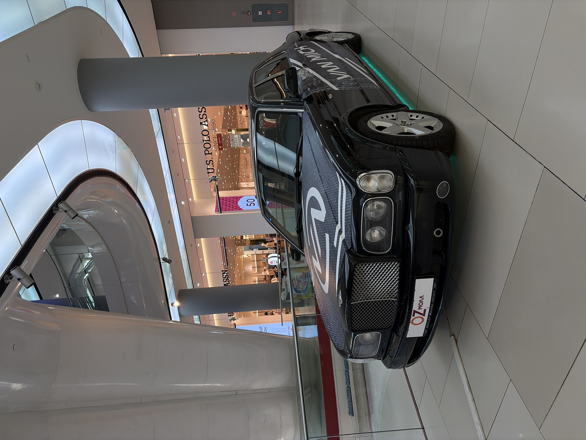 A black luxury sedan, likely a Bentley, is prominently displayed on a tiled floor inside a modern shopping mall. The car features custom white branding, including the text "VAN MICH" along its side and a large graphic on the hood. Green LED underglow illuminates the area beneath the vehicle.
The setting is a brightly lit indoor environment with contemporary architectural elements, including curved white ceilings with luminous panels, sturdy grey pillars, and glass railings on an upper level. No people are visible, but the presence of retail stores suggests an active commercial space.
Visible text includes a store sign for "U.S. POLO ASSN. SINCE 1890" and a "50%" sale banner nearby. The car's front license plate holder displays "OZ МОЛЛ" (OZ Mall) and "BENTLEY MOCKBA 933 3377" (Bentley Moscow 933 3377), indicating the car's origin or dealership, while the broader location context provided is Krasnodar, Russia. Security cameras are mounted on the architectural structures. The scene captures a static display within a busy commercial center, likely during daytime or evening hours given the artificial illumination.