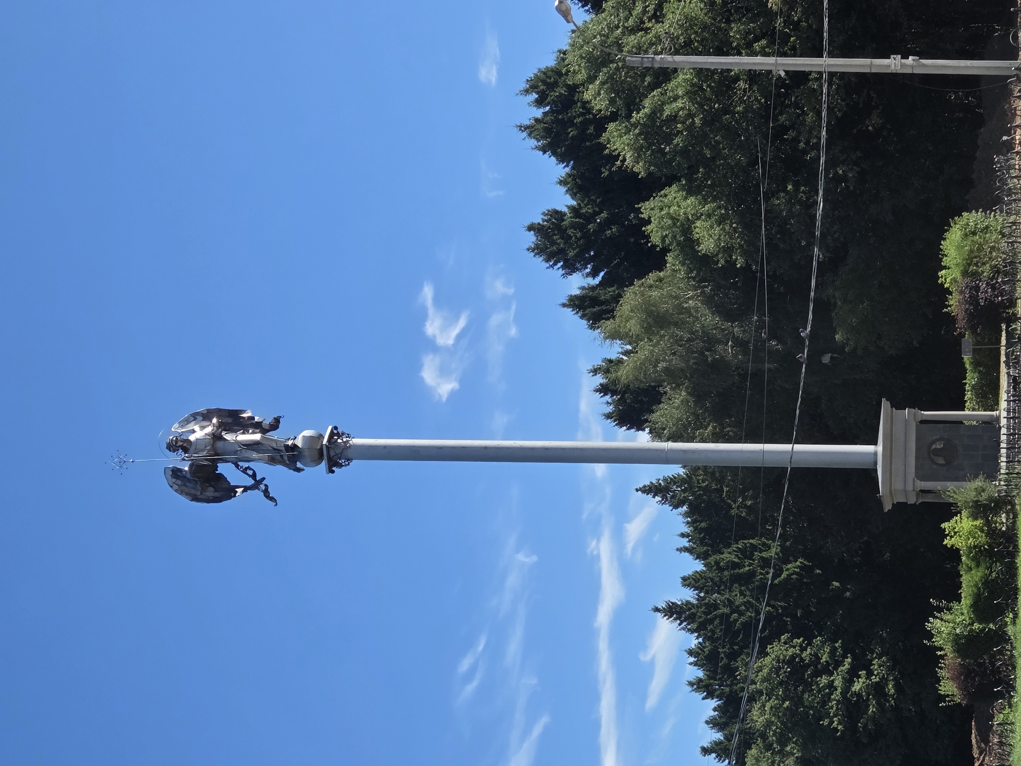 This image, taken in the city of Podolsk, Russia, depicts a tall monument topped with a statue of an angel. The angel, made of metal, is standing on a sphere and holds a staff topped with a cross in its right hand. The angel's wings are spread wide, and it appears to be looking downwards. The monument is situated on a grey, cylindrical pedestal, which rests on a square stone base with decorative carvings. Behind the monument, a dense forest of green trees forms a backdrop against a bright blue sky dotted with wispy white clouds. In the foreground, there is a patch of green grass and some low-lying bushes. A single utility pole stands to the right of the monument, with wires stretched across the scene. The overall impression is one of a serene, possibly historical, monument in a natural setting.