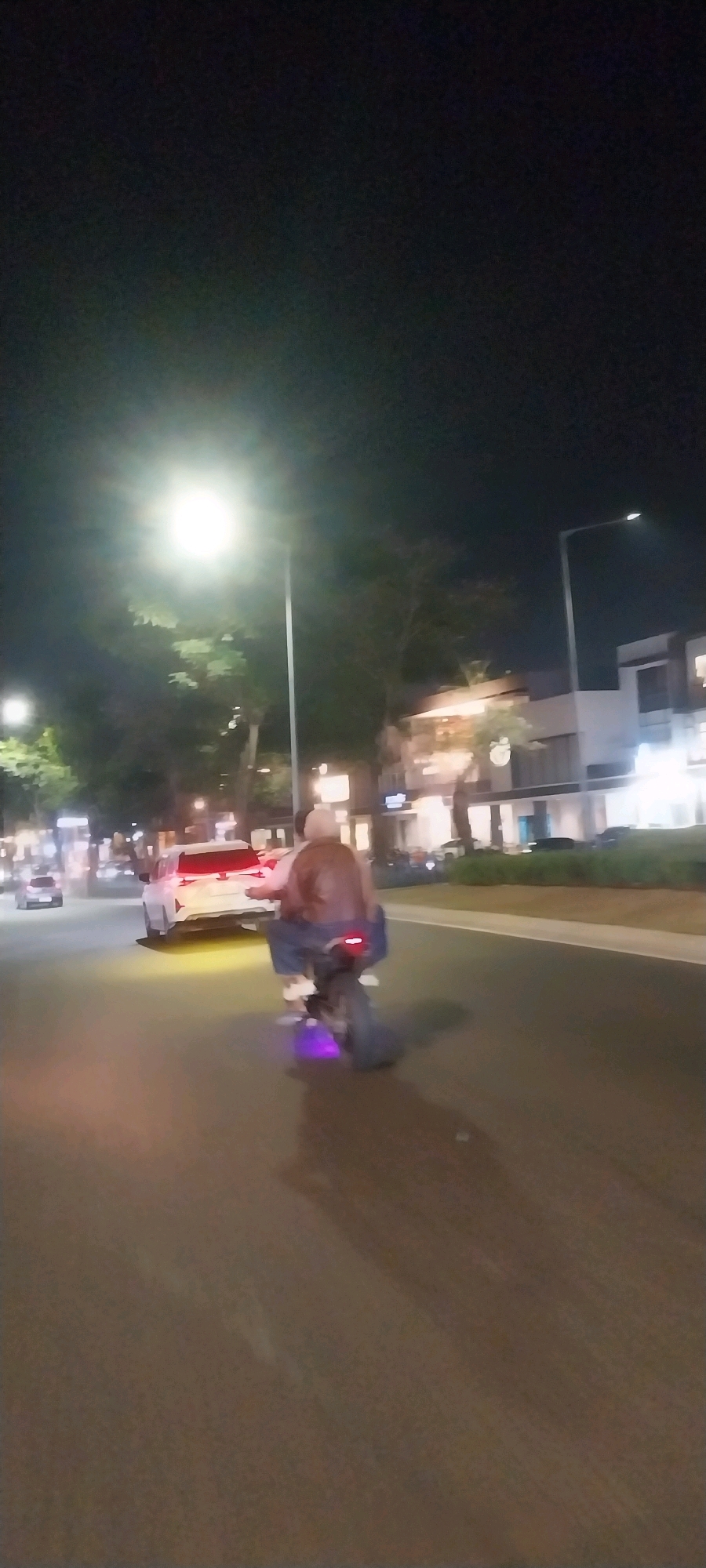At night, on a city street in Medan, Indonesia, a motorcycle with two people is captured in motion. The person in the front appears to be wearing a brown leather jacket and blue jeans, while the person behind them is dressed in a white shirt and blue jeans. The motorcycle has a distinct purple light emanating from its rear. In the background, a white car with illuminated red taillights is also present. The scene is illuminated by streetlights and the lights of buildings lining the street. The road surface is asphalt, and there are trees and sidewalks visible, suggesting an urban environment. The overall image has a low-light, slightly blurry quality due to the nighttime setting and motion.