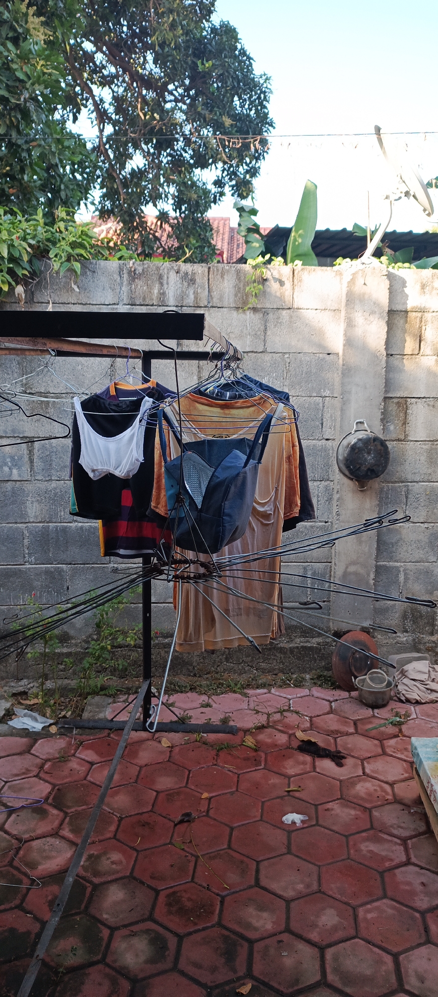 The image depicts an outdoor scene in Arjasa, Indonesia, featuring a clothesline with various garments hanging to dry against a textured gray cinder block wall. A metal drying rack stands prominently in the foreground, supporting shirts and other clothing items suspended on hangers. To the right of the rack, a dark pot is affixed to the wall. The ground is paved with hexagonal red tiles, some of which appear wet. Scattered debris, including leaves and a white plastic bag, is visible on the tiled surface. Lush green foliage and trees are present in the background, and the sky is a clear blue. The overall impression is one of a typical domestic setting where laundry is being air-dried.