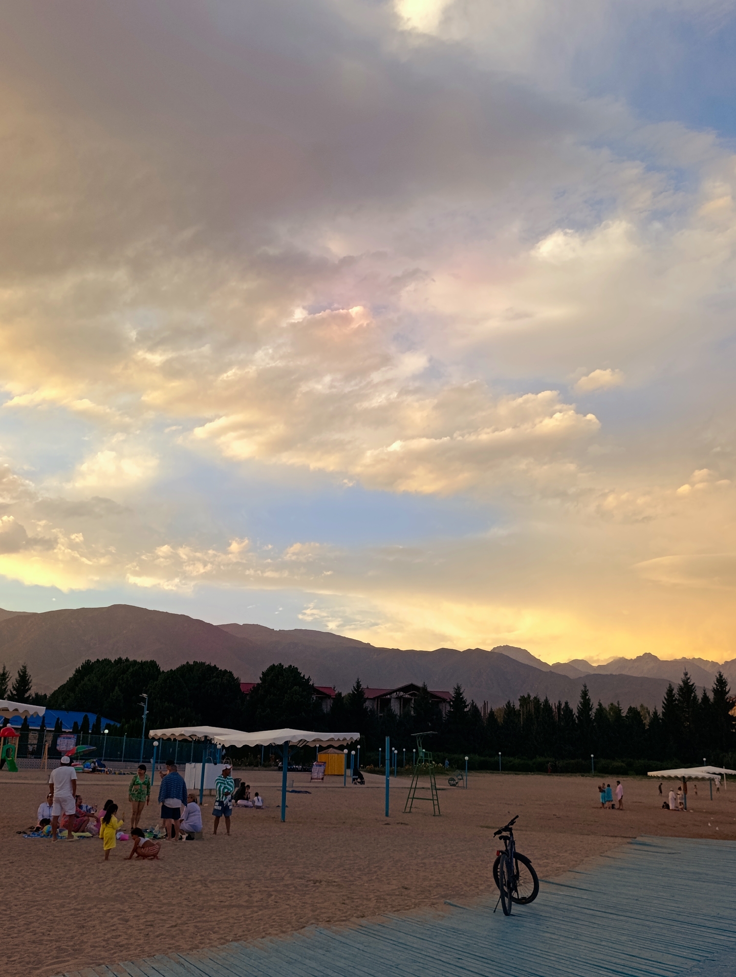 The image depicts a sandy beach area with people gathered, set against a backdrop of mountains and a dramatic sky during what appears to be sunset. The location is identified as Bayet, Kyrgyzstan.
In the foreground, a wooden walkway made of light-colored planks with blue painted edges is visible. A dark-colored bicycle with a red reflector is parked on the sand near the walkway.
The middle ground of the image shows a beach populated by several individuals and groups. Some people are sitting on the sand, while others are standing. There are white canopy structures providing shade. In the background of this area, there are trees, buildings with red roofs, and a playground structure with colorful slides and a ladder.
The far background features a range of mountains, their peaks silhouetted against the sky. The sky is filled with clouds, illuminated by warm hues of orange, yellow, and pink, suggesting the sun is setting or has recently set. Some clouds are darker gray.
The overall impression is a lively recreational scene on a beach in a mountainous region.