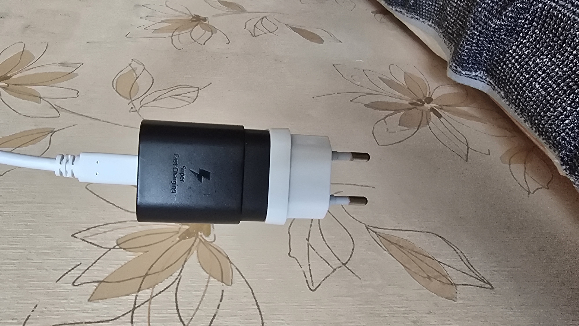 A close-up, vertical shot shows a white USB-C cable connected to a black Samsung fast-charging adapter, which is then plugged into a white European-style power adapter with two prongs. The adapter is suspended against a light beige wallpaper with a subtle floral pattern in light brown and white. The background wallpaper exhibits a repeating design of stylized flowers and leaves. The image is devoid of any people or identifiable landmarks in Novohuivynske, Ukraine. The lighting is moderate, casting soft shadows and highlighting the textures of the wallpaper and the plastic adapters.