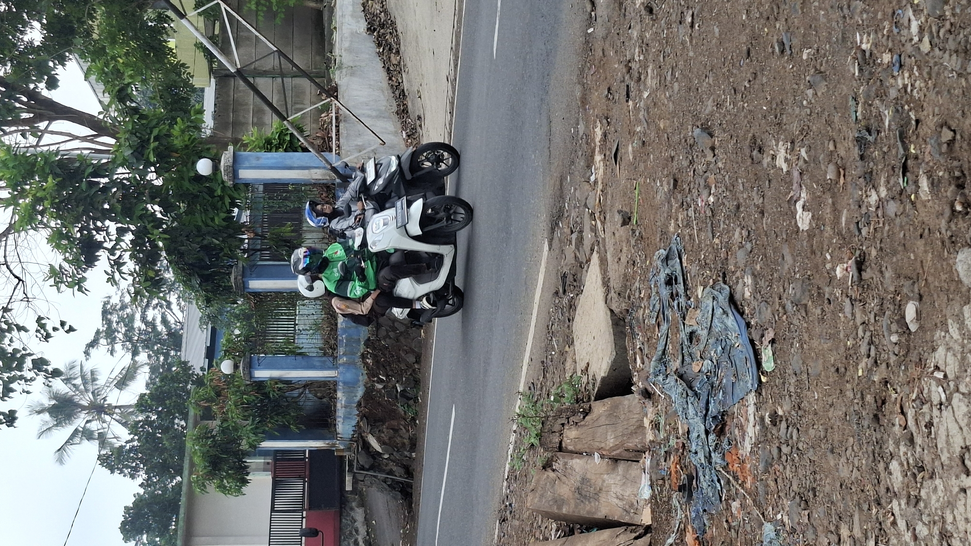 Two people ride on a white scooter, with the driver wearing a green jacket and a helmet, and the passenger behind wearing a blue helmet and a grey jacket. Another person rides a black scooter behind them, wearing a blue helmet and a grey jacket. They are on a paved road with a white line dividing the lanes. To the left of the road is a blue and white fence with a building behind it. To the right of the road is a dirt area with scattered debris, including some large wooden planks and blue tarp. The scene appears to be in Cilaku, Indonesia.
