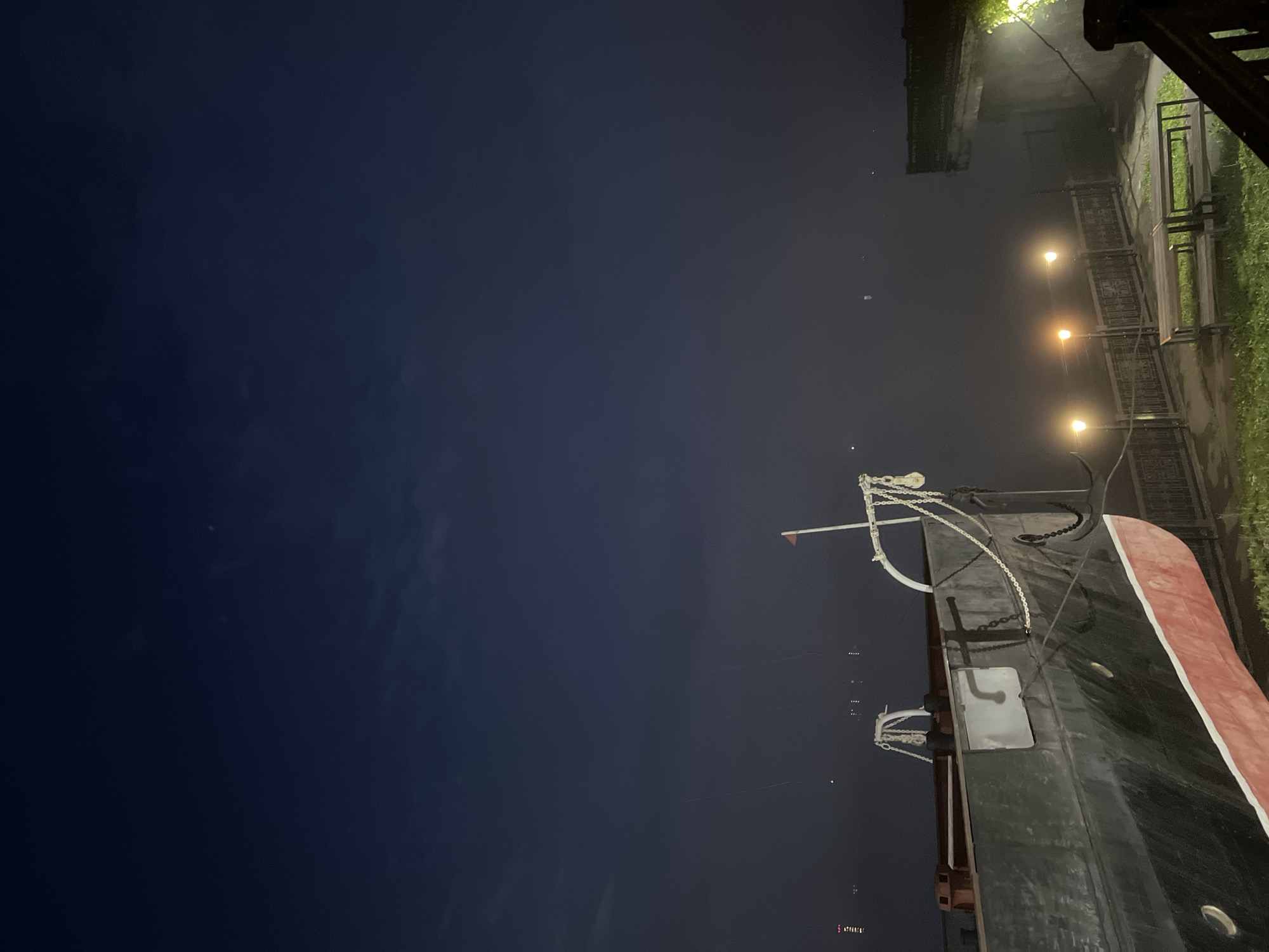 The image depicts a moored ship at night, likely in Krasnoyarsk, Russia, given the provided context. The ship, primarily black with a red waterline, is the dominant subject in the foreground, angled from the lower left towards the center. Its bow is visible, adorned with chains and a pulley system. A small red flag flies from a white pole at the ship's stern.
In the middle ground, a waterfront promenade is suggested by a metal railing and several lampposts casting warm yellow light. Two empty wooden benches are positioned on the grass beside the promenade. Beyond the promenade, the dark water of a river or bay is visible, with faint lights from the opposite shore or other vessels appearing as distant smudges.
The sky is a deep, dark blue, with a few wispy clouds. The overall atmosphere is calm and quiet, characteristic of a nighttime scene in a waterfront area. There are no people or animals visible in the frame. The scene appears to be a public space, possibly a park or a promenade adjacent to a pier.