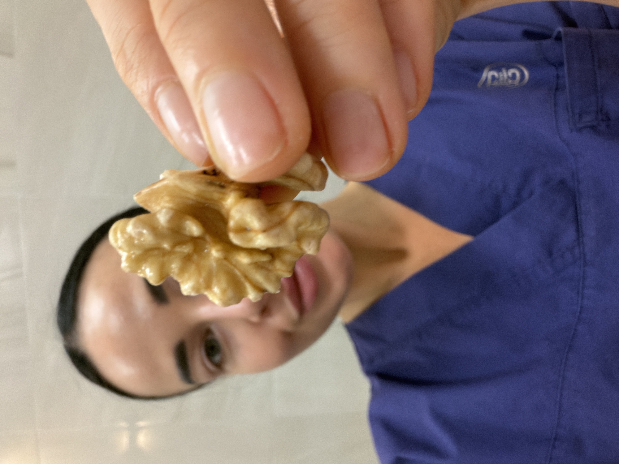 A person holds a shelled walnut in their fingers. The person has fair skin, dark hair, and is wearing a blue garment with an embroidered logo on the upper right chest. The background includes a blurred face and a light-colored wall. Location cannot be determined.