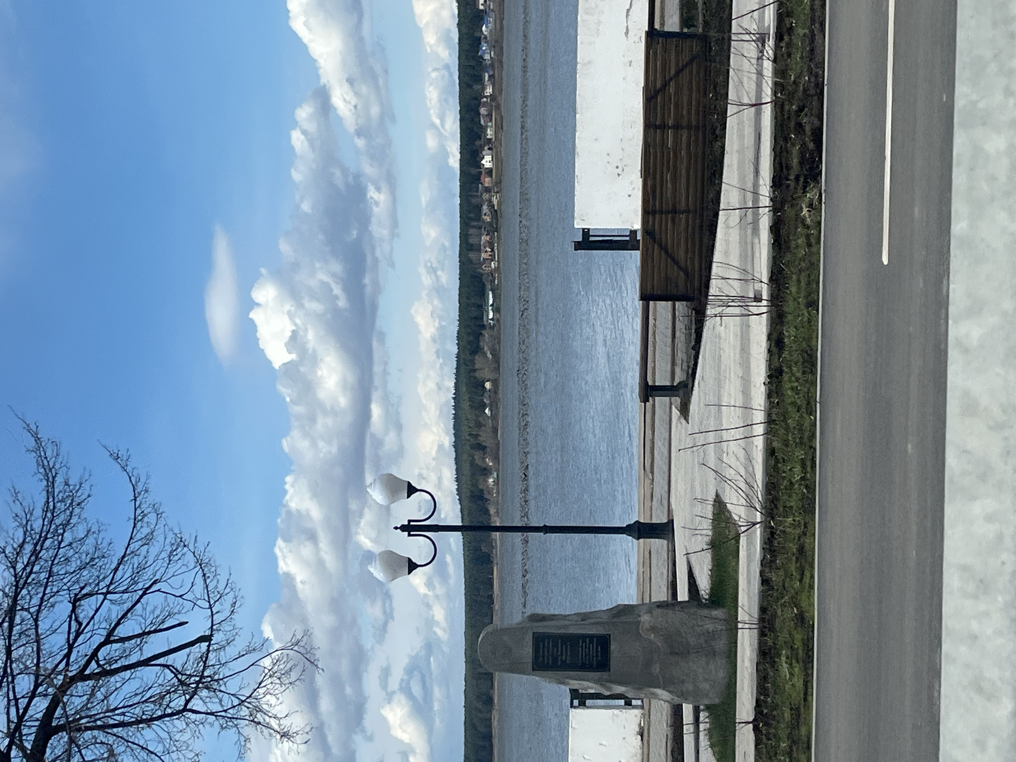 The image displays a serene waterfront promenade in Votkinsk, Russia, under a bright blue sky adorned with scattered white clouds. A wide body of water, likely a river or lake, stretches across the midground, its surface gently reflecting the sky. On the distant shore, a continuous line of dense evergreen trees is visible, with scattered small buildings or houses nestled within, suggesting a quiet settlement.
In the foreground, a paved road or pathway with a white lane marking runs alongside a grassy verge, which in turn borders a paved pedestrian area. To the left, a bare tree with intricate, leafless branches stands out against the sky, indicating a late winter or early spring season. A black, ornamental lamppost with two white, opaque globes provides illumination for the promenade. Adjacent to the lamppost is a stone monument or pedestal featuring a dark, rectangular plaque. While the text on the plaque is too small to discern, it appears to be in Cyrillic, consistent with the Russian location.
Further along the promenade to the right, a wooden slatted bench faces the water, suggesting a spot for relaxation. Nearby are parts of a white building or structure and a dark fence. The scene is devoid of people or any specific activities, conveying a sense of peace and stillness. The bright natural light indicates it is daytime, likely a sunny morning or afternoon, with pleasant weather conditions.
