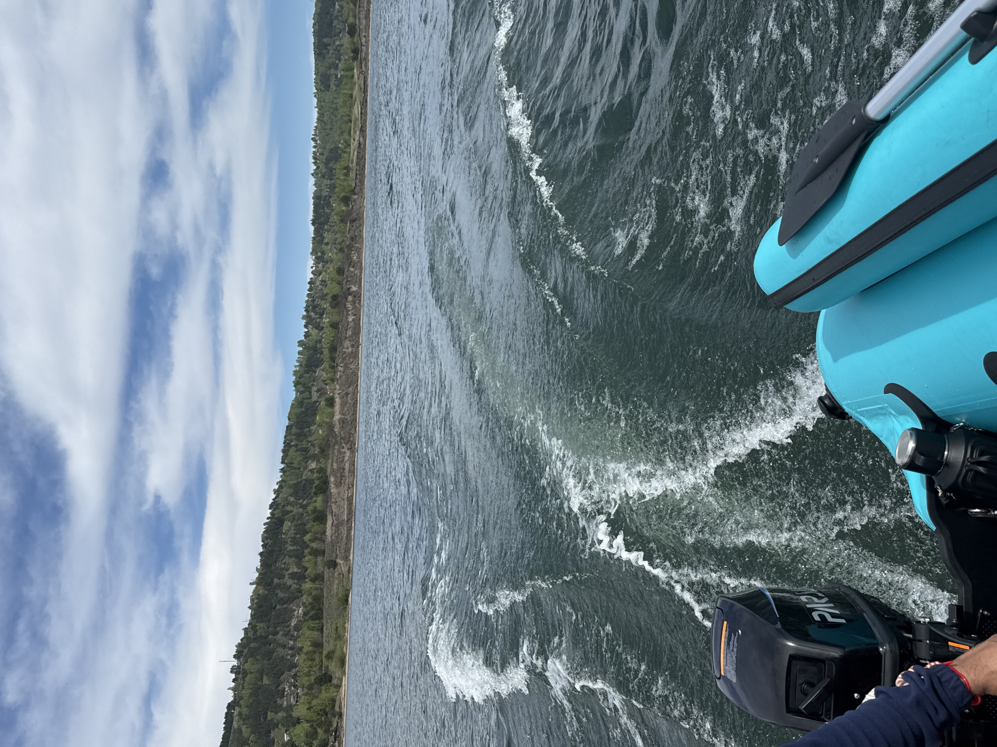 The image depicts a view from a light blue inflatable boat on what appears to be a body of water, possibly a lake or river. The boat is equipped with a black outboard motor and a paddle rests on its side. The water is dark green with white wake trailing behind the boat. On the left side of the frame, the shoreline is visible, covered with dense green trees and some areas of exposed rock. The sky above is light blue with patches of white clouds. No people are clearly visible, although part of a hand and sleeve can be seen near the motor.
Given the information, it's challenging to definitively pinpoint the exact location in Irkutsk, Russia. However, Irkutsk is near Lake Baikal, so it is plausible that the scene depicts the Angara River, or a smaller body of water in the vicinity. The presence of the shoreline and the type of vegetation would be consistent with such an environment.