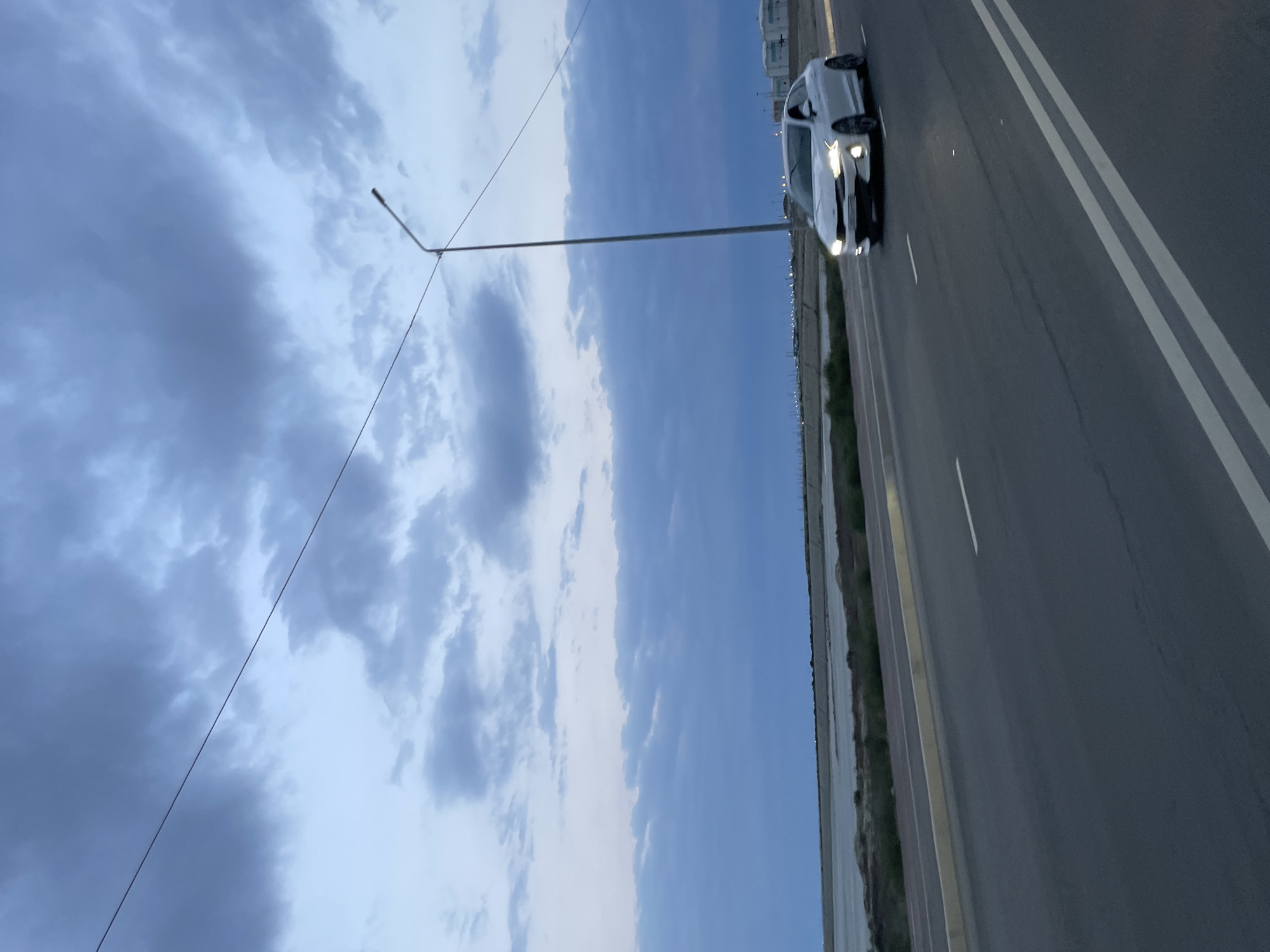 A white sedan drives on a multi-lane road in Ömirzaq, Kazakhstan. The car's headlights are on, illuminating the road ahead. The sky above is overcast with dramatic clouds in shades of blue and gray. A tall lamppost stands to the left of the road, with a wire stretching diagonally across the frame. To the right of the road is a body of water, possibly a lagoon or a lake, with a flat, man-made embankment. In the distance, faint structures and lights suggest the presence of a town or industrial area. The overall scene depicts an outdoor environment during twilight hours, characterized by a muted color palette and atmospheric lighting.