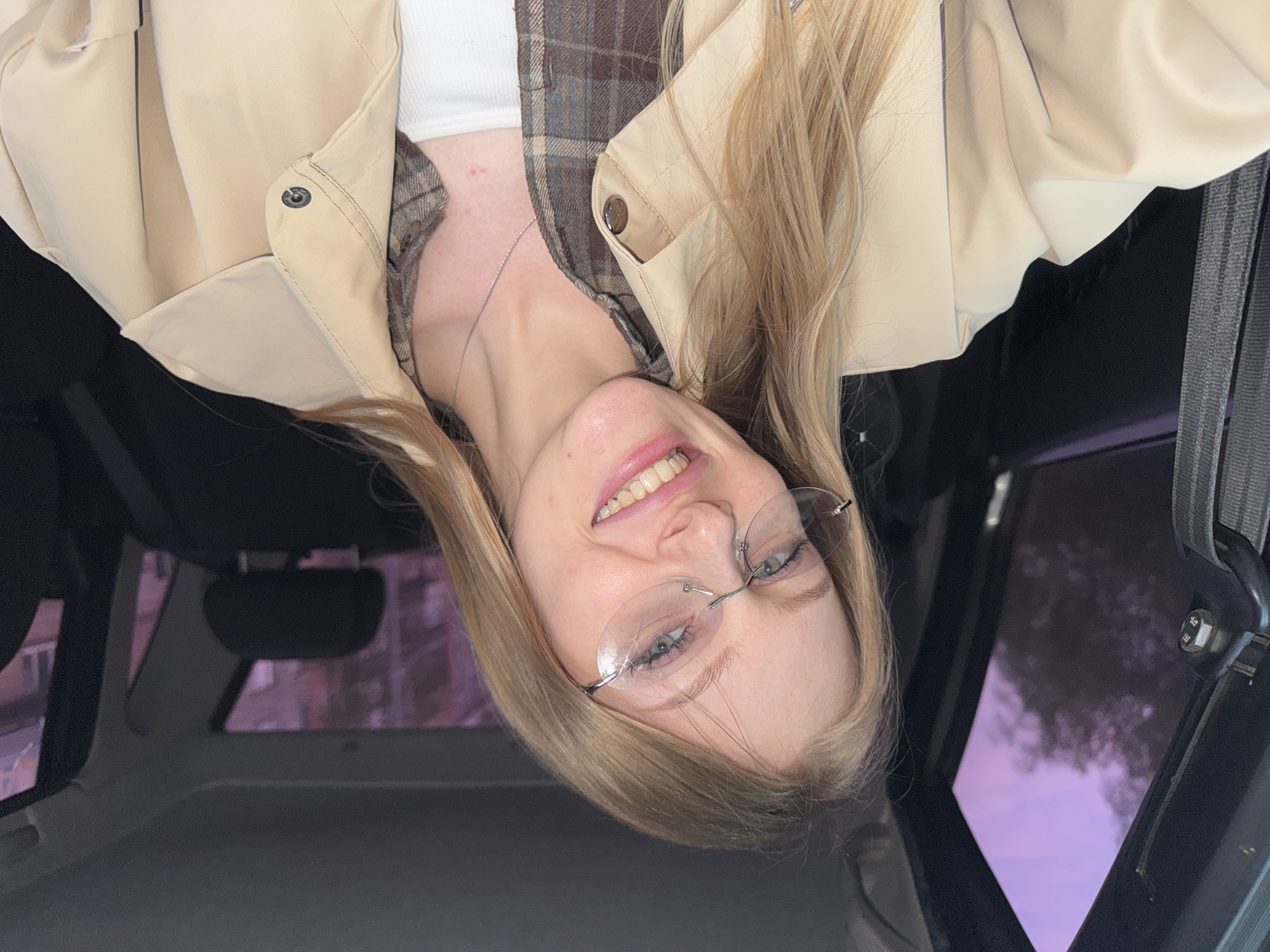 A woman with blonde hair is inside a car, wearing glasses, a white top, a plaid shirt, and a beige jacket. The background shows a car interior and a glimpse of a city through the sunroof. The city location is indicated as Perm, Russia.