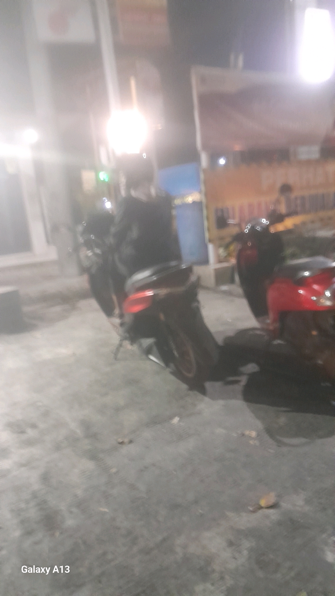 The image shows a blurry outdoor scene at night, likely in Jekulo, Indonesia, based on the text "Galaxy A13" at the bottom left. Two motorcycles are visible. The motorcycle in the foreground is a dark-colored scooter, partially obscuring a red scooter behind it. A person wearing a dark jacket is seated on the dark scooter, facing away from the camera. Behind the motorcycles, a brightly lit object, possibly a lamp or light fixture, casts a strong glow, creating lens flare. In the background, there are blurry structures and possibly a banner or sign with text that is illegible due to the blurriness and lighting conditions. The ground appears to be paved, with some debris scattered on it.