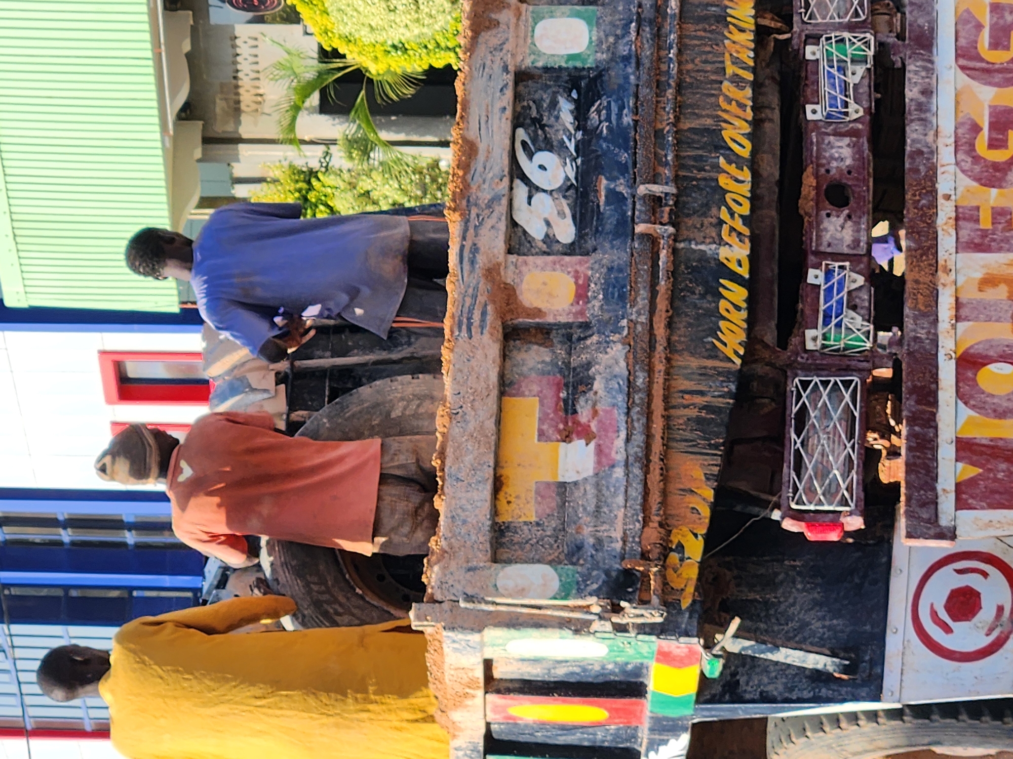 In an outdoor urban environment, three individuals are positioned on the back of a dirty truck. The person on the left wears a yellow robe, while the middle person is dressed in an orange shirt and a beanie. The person on the right is in a blue t-shirt and shorts. The truck's rear, which is heavily soiled with mud and paint, features various colorful markings, including numbers, a yellow "T" shape, and a prominent yellow inscription that reads "HORN BEFORE OVERTAKING." The truck has large tires visible on the left side. In the background, a building with blue and white vertical panels and a green corrugated roof can be seen.
