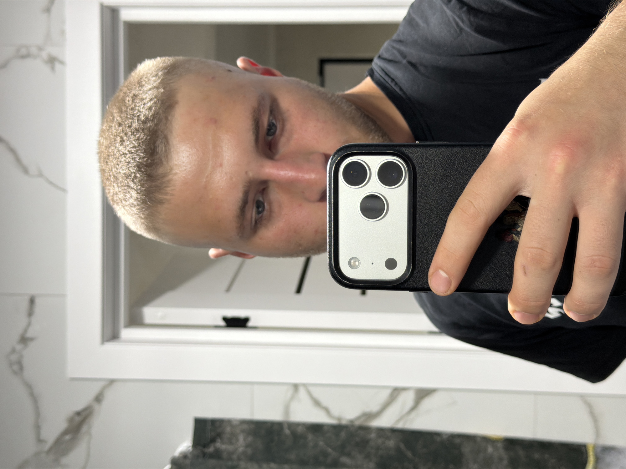 A young Caucasian man with a buzz cut is taking a selfie in a bathroom. He is holding a black phone with a silver camera module in his right hand, angled towards the mirror. The man is wearing a black t-shirt. The bathroom features white trim around a doorway, marble-patterned tiles on the wall, and a dark countertop with a mirror reflecting part of the room. The lighting suggests an indoor setting. The image is taken from a low angle, focusing on the man's face and the phone.