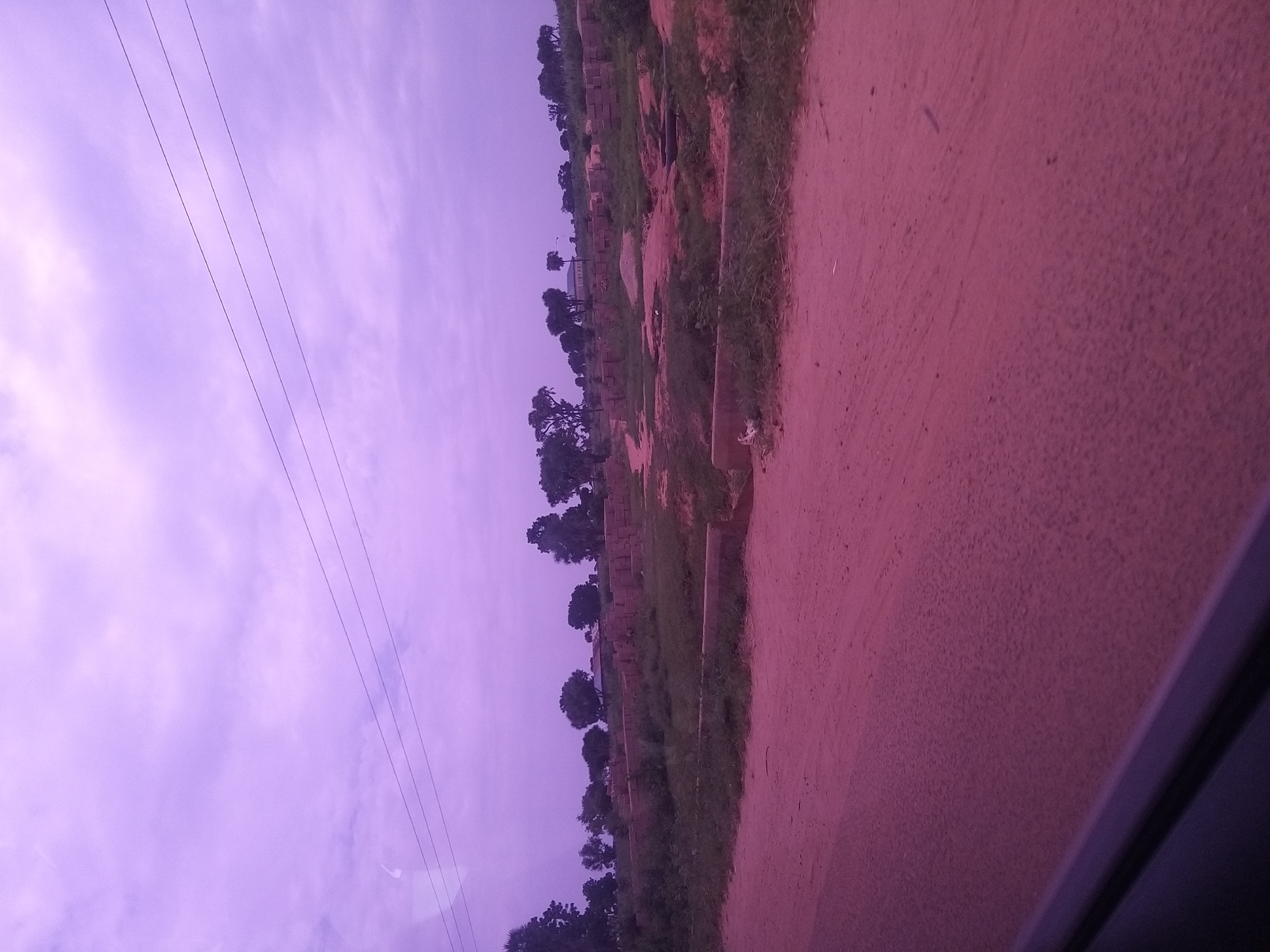 The image captures a scene in Kafarati, Nigeria, under a hazy, purple-tinged sky. A dirt road, with a reddish hue, dominates the foreground, suggesting a rural or undeveloped area. In the mid-ground, a construction site is visible, with stacks of bricks and building materials scattered across uneven terrain. Patches of grass and low vegetation grow between the construction elements. Several trees, some appearing mature and others saplings, dot the landscape behind the construction area. In the distance, a building, possibly a house or a small commercial structure, can be seen nestled among the trees. Three thin, dark power lines stretch horizontally across the upper portion of the sky, indicating the presence of electrical infrastructure. The overall impression is one of ongoing development or a partially completed project in a semi-arid environment.