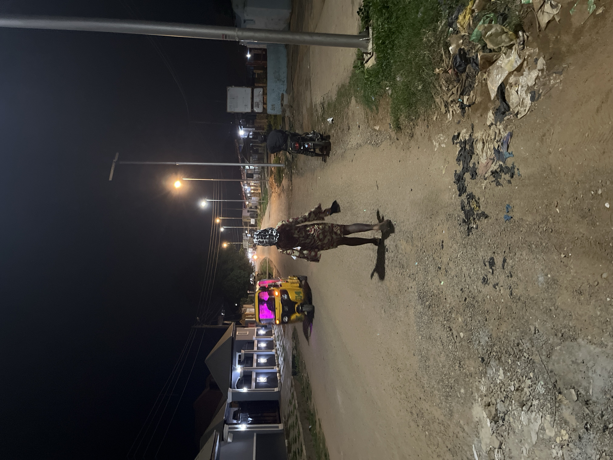 The image captures a night scene on a dirt road in Abuja, Nigeria. In the foreground, a person walks away from the camera, wearing a patterned dress and a headscarf. To the left, a yellow auto-rickshaw, known as a 'Keke Napep' in Nigeria, is parked, emitting a pink glow from its lights. Further down the road, a person is seated on a motorcycle, facing away from the camera. The street is lined with houses and streetlights, some of which are illuminated. The road surface is unpaved and shows signs of wear and tear. Piles of trash are visible on the right side of the road. The overall impression is of a quiet, dimly lit street in a residential area.