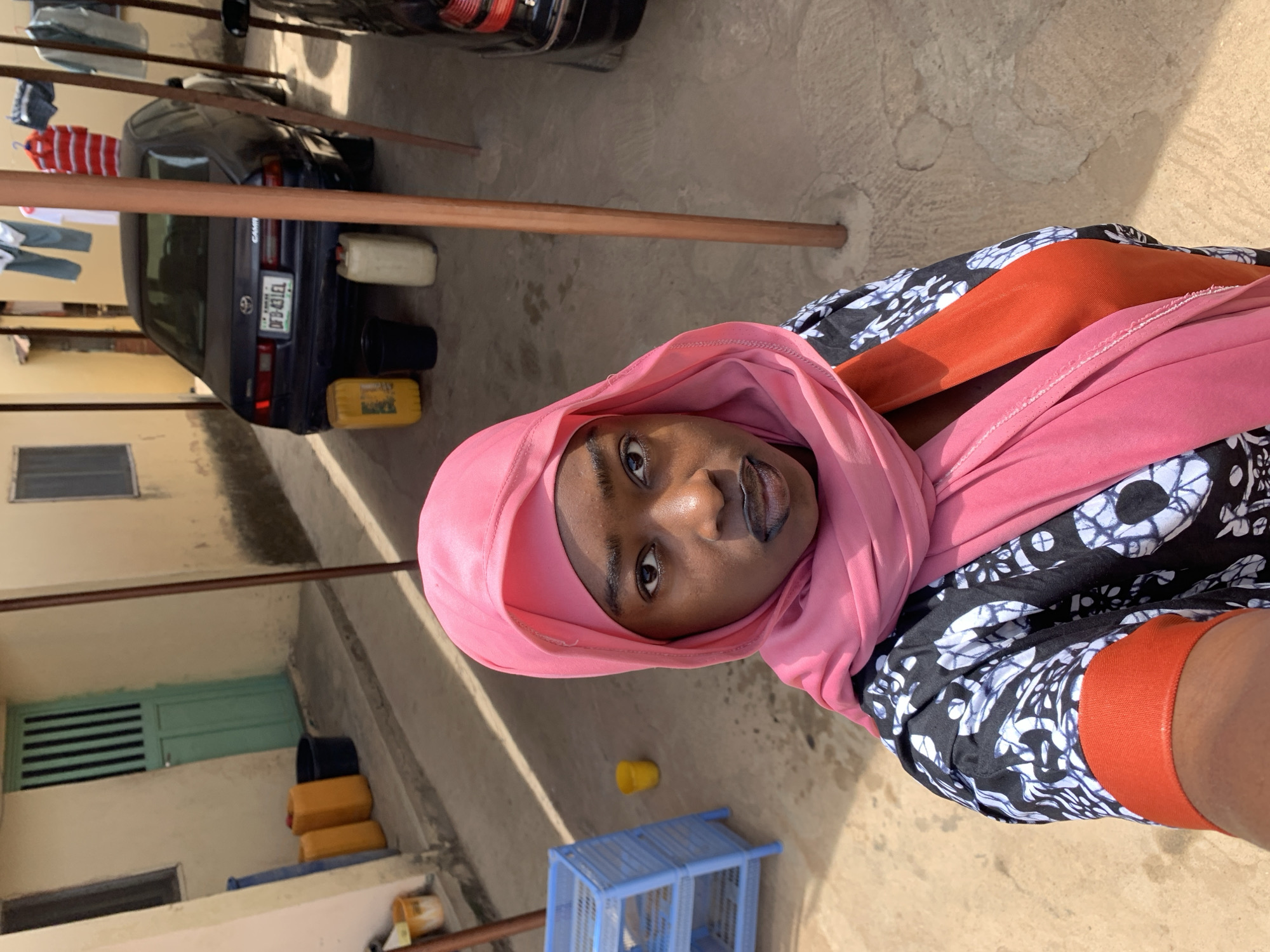 A young woman in Rafin Par, Nigeria is seen from a low angle. She is wearing a pink hijab, an orange top, and a black-and-white patterned dress. She has dark skin and is looking directly at the camera. Behind her are buildings, a car, and clothes hanging on a line. The buildings are light-colored, and the car is dark-colored. The ground is a light-colored concrete.