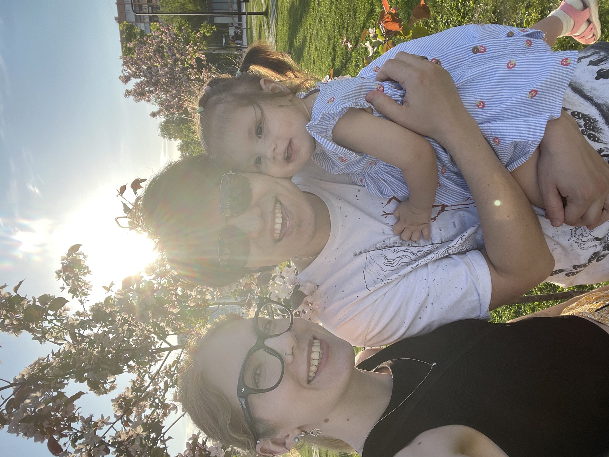 The image shows three people outdoors in a sunny setting. A woman wearing glasses and a black top is at the bottom of the frame. Above her is another adult wearing sunglasses and a white t-shirt, holding a young child in a blue and white striped dress with strawberry patterns. The child is looking towards the camera. The background includes a grassy area, trees, and blooming branches. The bright sun is shining through the trees, causing lens flare. Based on the location, it is possible that this photo was taken in Izhevsk, Russia, although no specific landmarks are visible to confirm this.