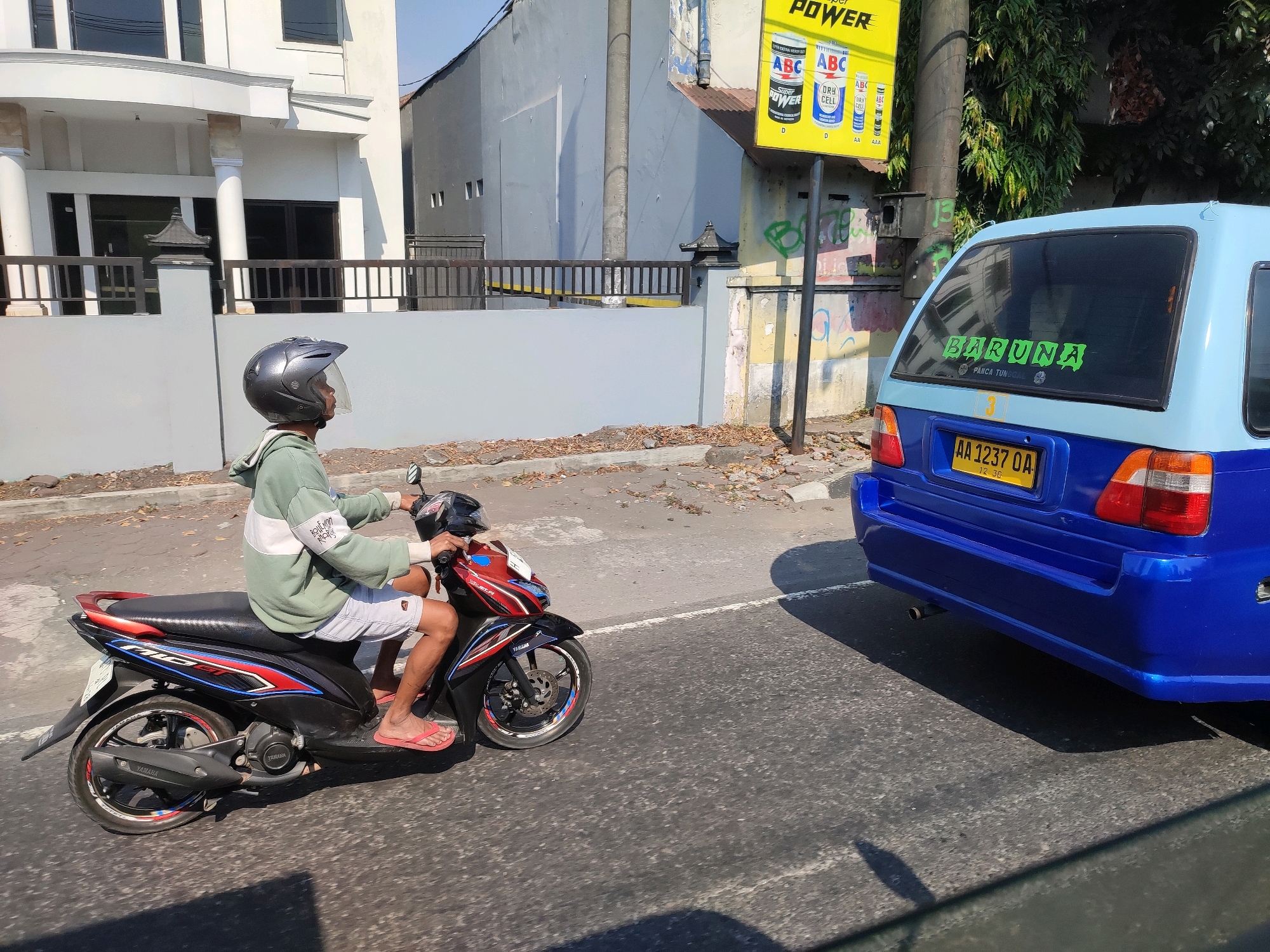 In Magelang, Indonesia, a person wearing a dark gray helmet and a green and white hooded jacket rides a black, red, and blue motorcycle. The person is also wearing white shorts and red flip-flops. The motorcycle has a license plate reading "AA 1237 QA". To the right of the motorcycle is the rear of a blue van with a green sticker on the back window that says "BARUNA". The van has a yellow license plate. The scene takes place on a paved road with some debris and dry leaves on the side. Behind the road, there is a white building with columns and a fence, and further back, a grey building. A yellow billboard with advertisements for "POWER" batteries and "ABC SUPER DRY CELL" is visible on the right side of the image. The sky is clear and blue, indicating a sunny day.