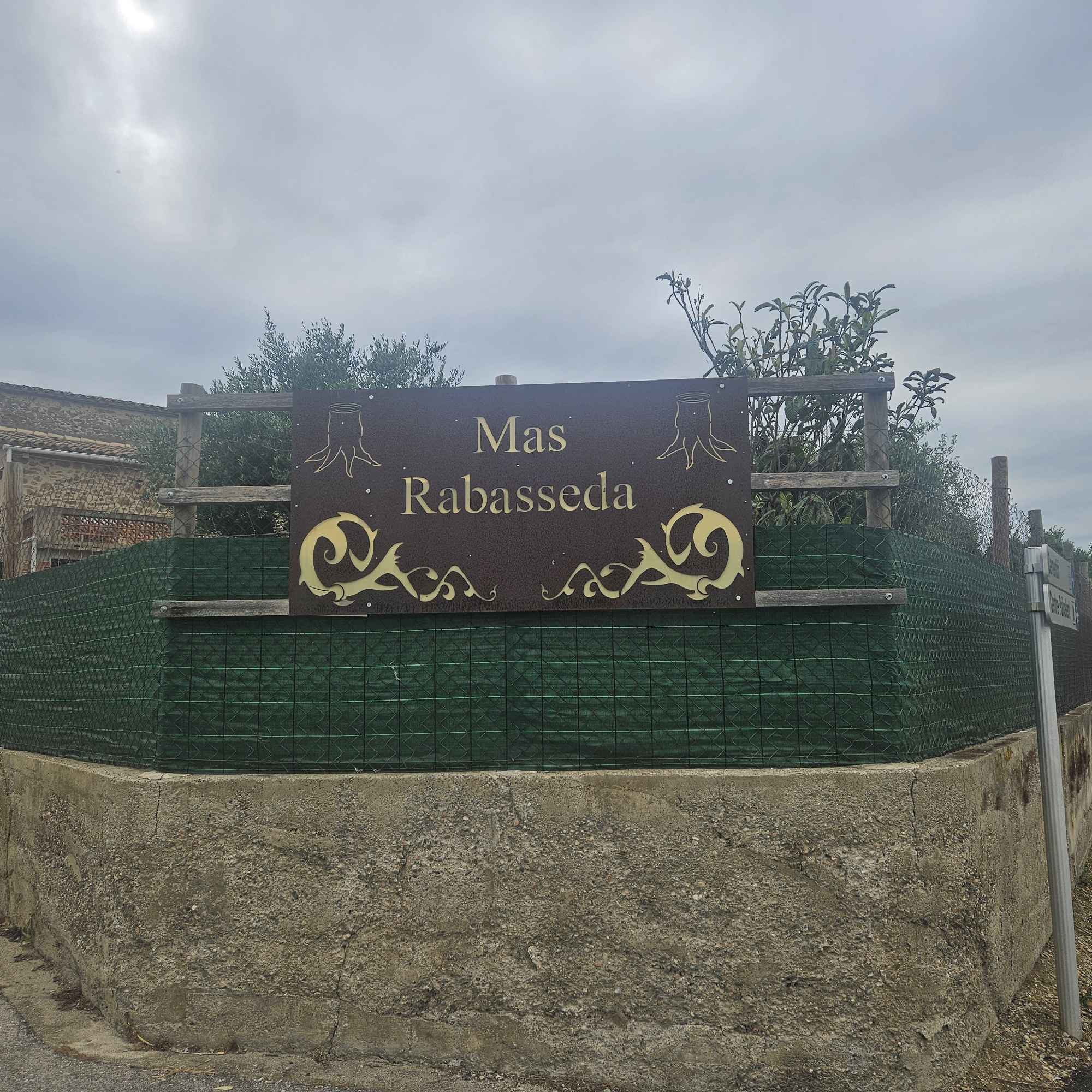 In Aiguaviva, Spain, a weathered sign with gold lettering on a dark brown, rusty metal background reads "Mas Rabasseda". The sign is mounted on wooden posts, part of a fence enclosure. To the left of the sign, a building with a stone facade and terracotta roof tiles is visible. The fence itself is made of green netting stretched over a wooden frame, supported by concrete. The ground in front of the fence is a rough, textured concrete wall. A street sign is visible on the right side of the image. The sky is overcast.