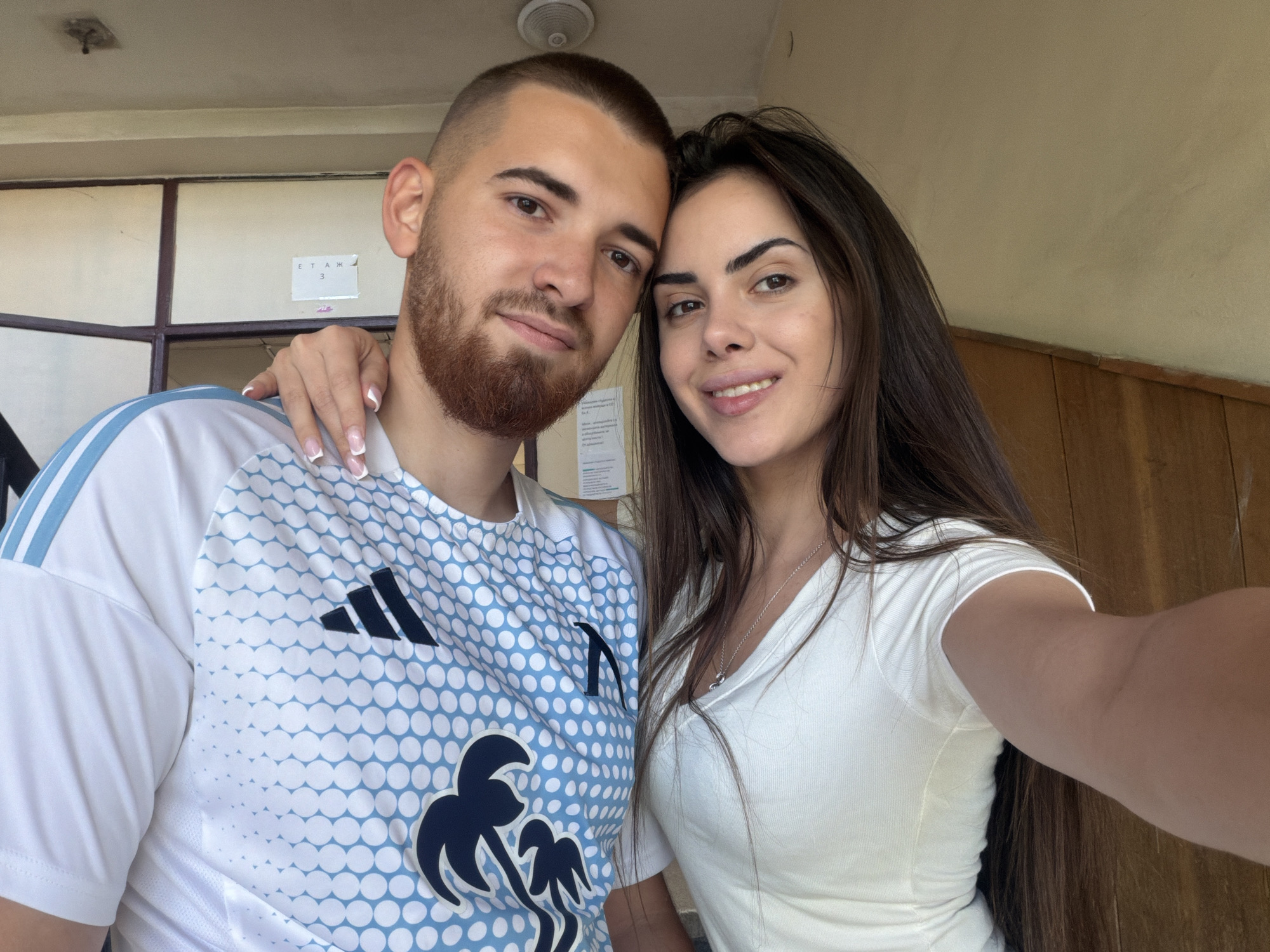 A selfie shows a light-skinned male and female, possibly a couple. The male has a shaved head with short brown hair and a full beard. He wears a white t-shirt with light blue dots and a dark blue design, potentially a sports jersey. The female has long brown hair and wears a white top. Her left hand is on the male's shoulder. A ceiling light fixture is visible in the upper center of the frame. A wall painted a light color and brown wooden paneling are in the background, along with a white sign with unreadable text. The location is possibly in Sofia, Bulgaria.
