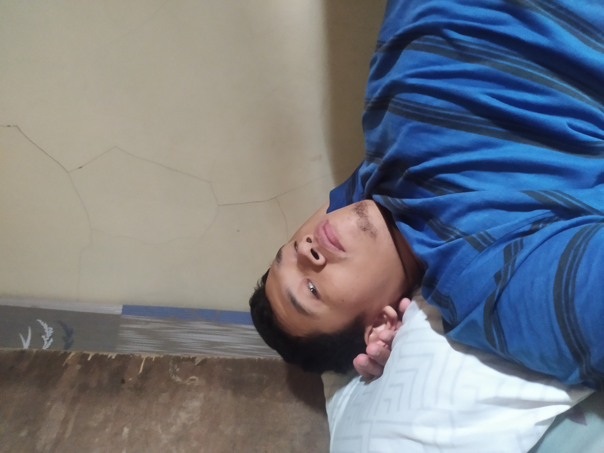 A young man is lying on his back on a pillow, looking upwards. He is wearing a blue t-shirt with black stripes. The background appears to be a wall with some cracks. The man's left hand is resting near his head. This image was taken indoors in the city of Kragilan, Indonesia.